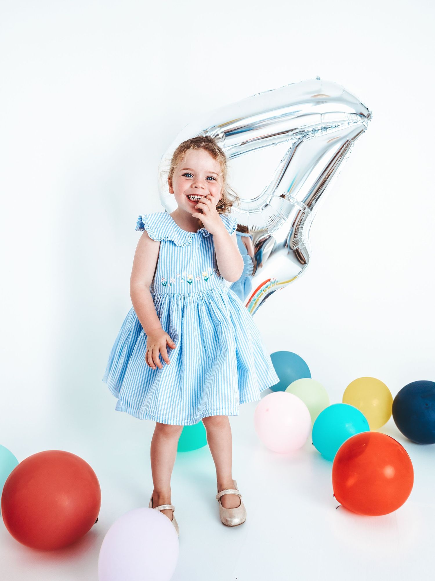 A smiling toddler girl in a blue dress celebrates her 4th birthday with colorful balloons and a silver number 4 balloon.
