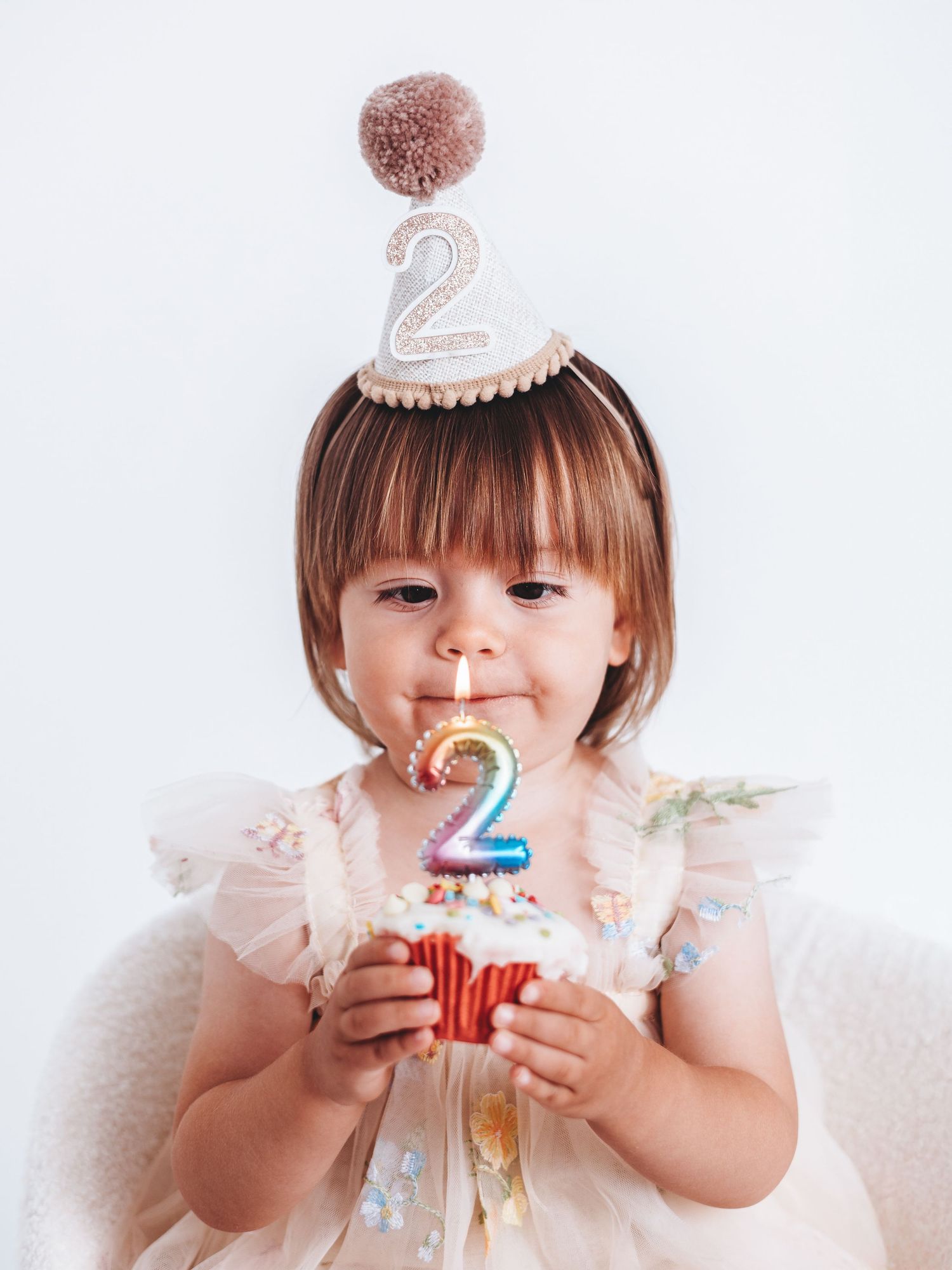 Toddler girl wearing a number 2 birthday hat holds a cupcake with a colorful number 2 candle, celebrating her 2nd birthday.