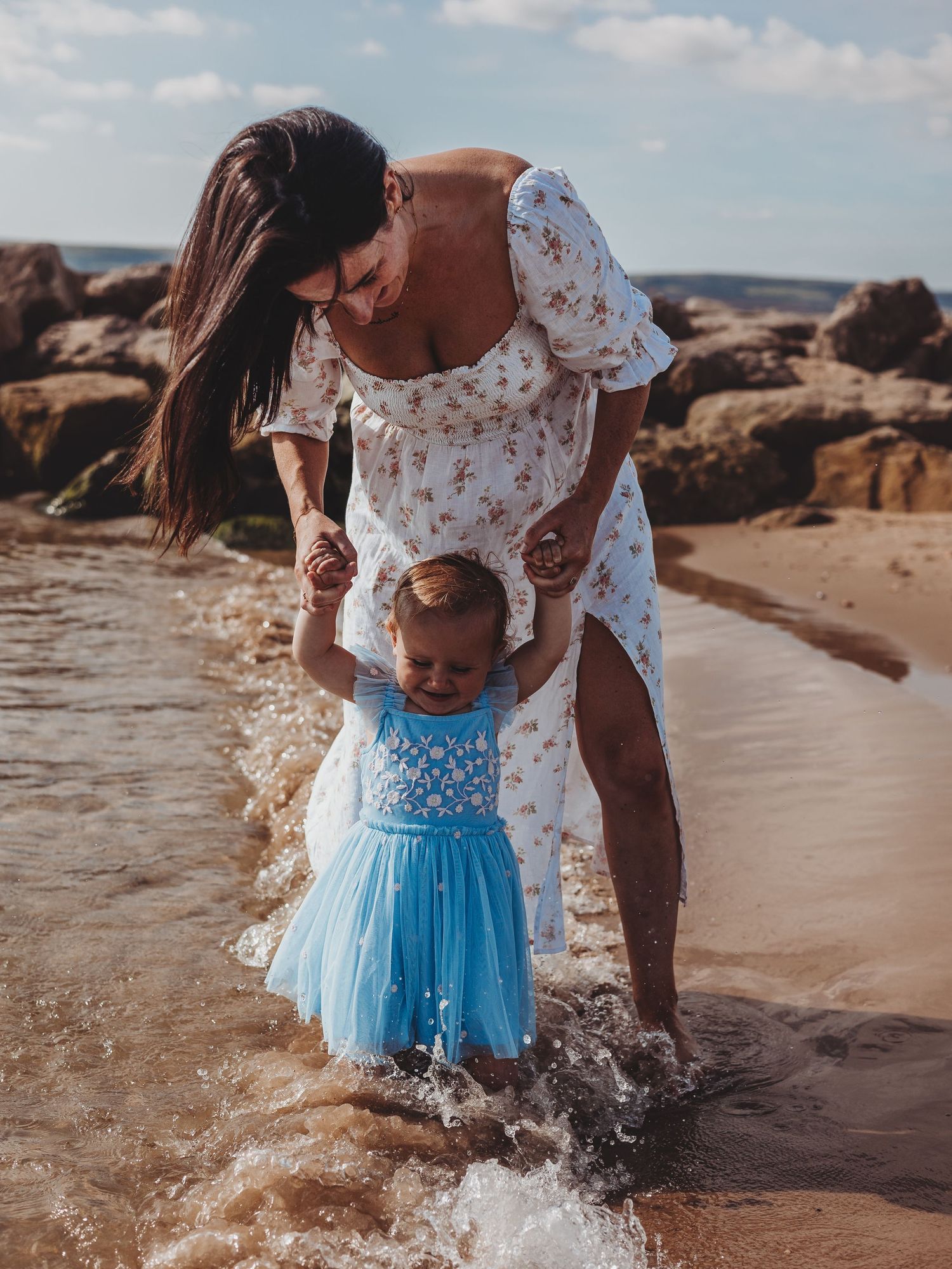 A mother in a floral dress helps her toddler girl in a blue tutu walk through shallow beach waves.