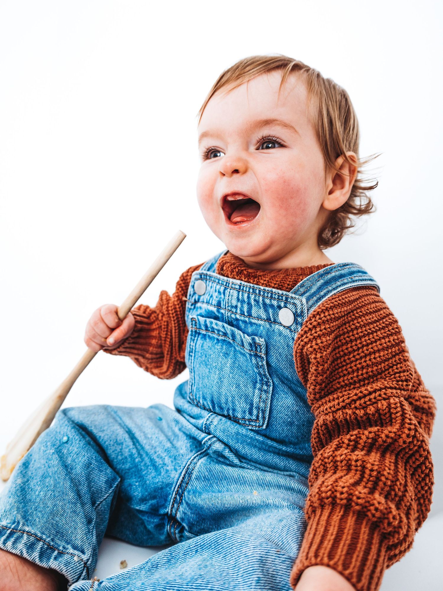 Happy toddler in denim overalls and rust knit sweater laughing while holding a wooden stick.