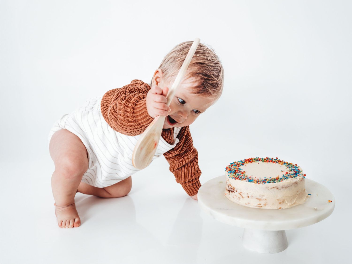 Baby in brown cardigan reaching for a colorful sprinkle cake on a white cake stand during first birthday smash.