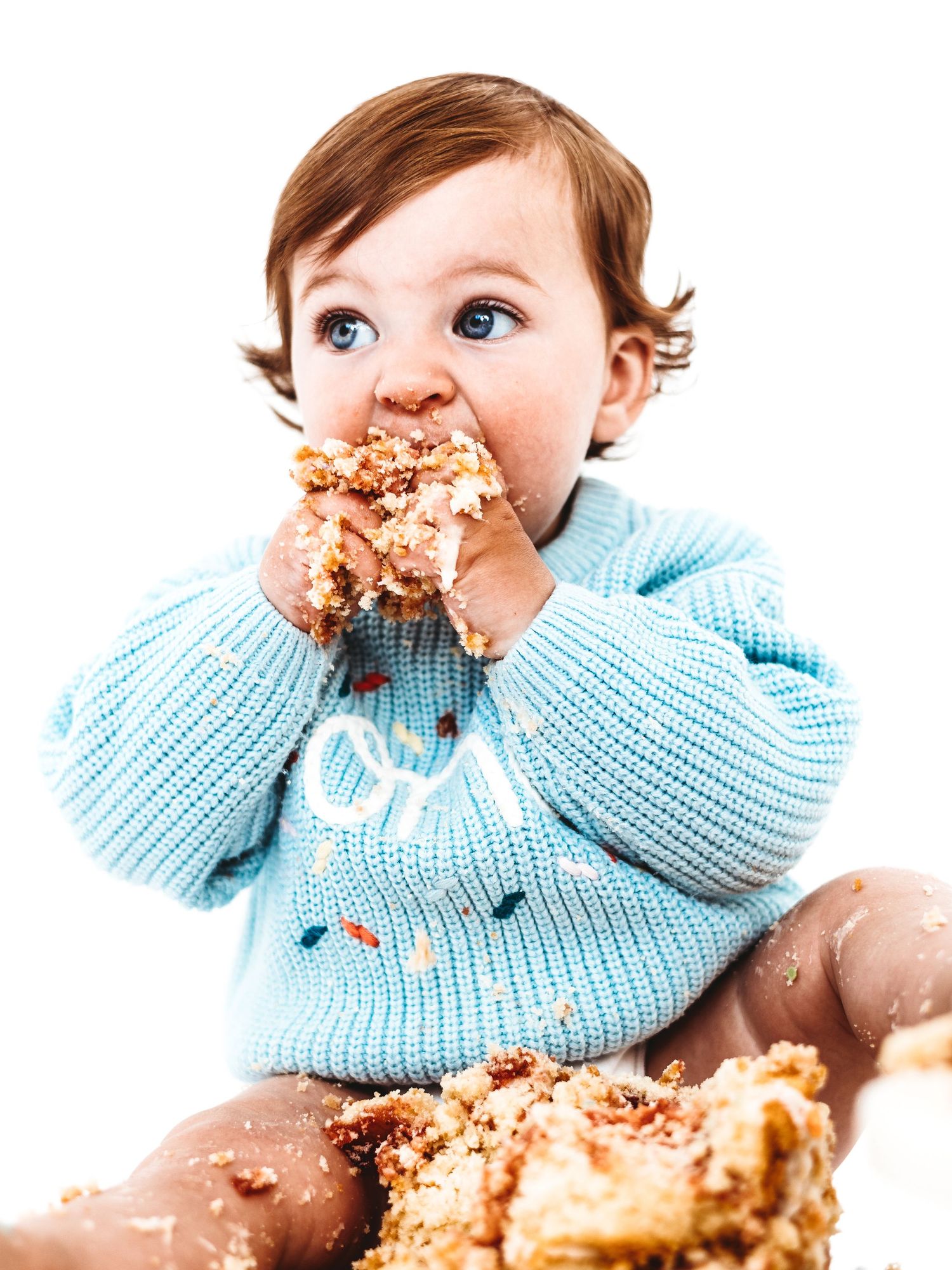 Messy toddler in blue sweater eating birthday cake with both hands, face covered in crumbs.