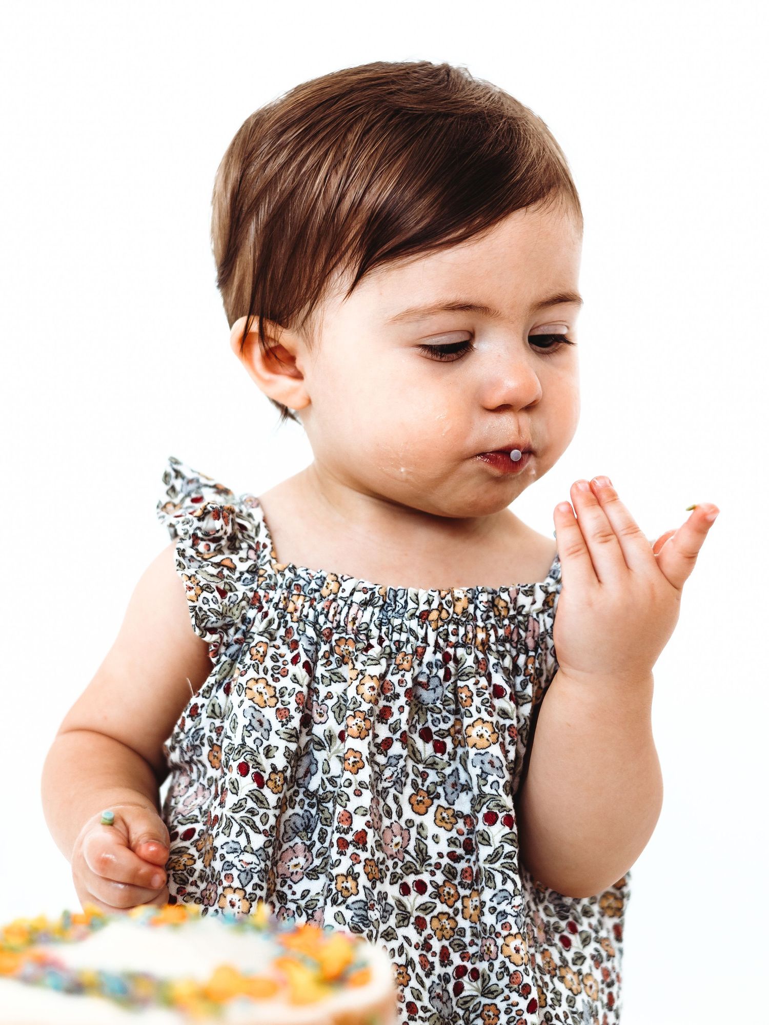 A baby girl in a floral dress tastes frosting at her first birthday cake smash on a white background.
