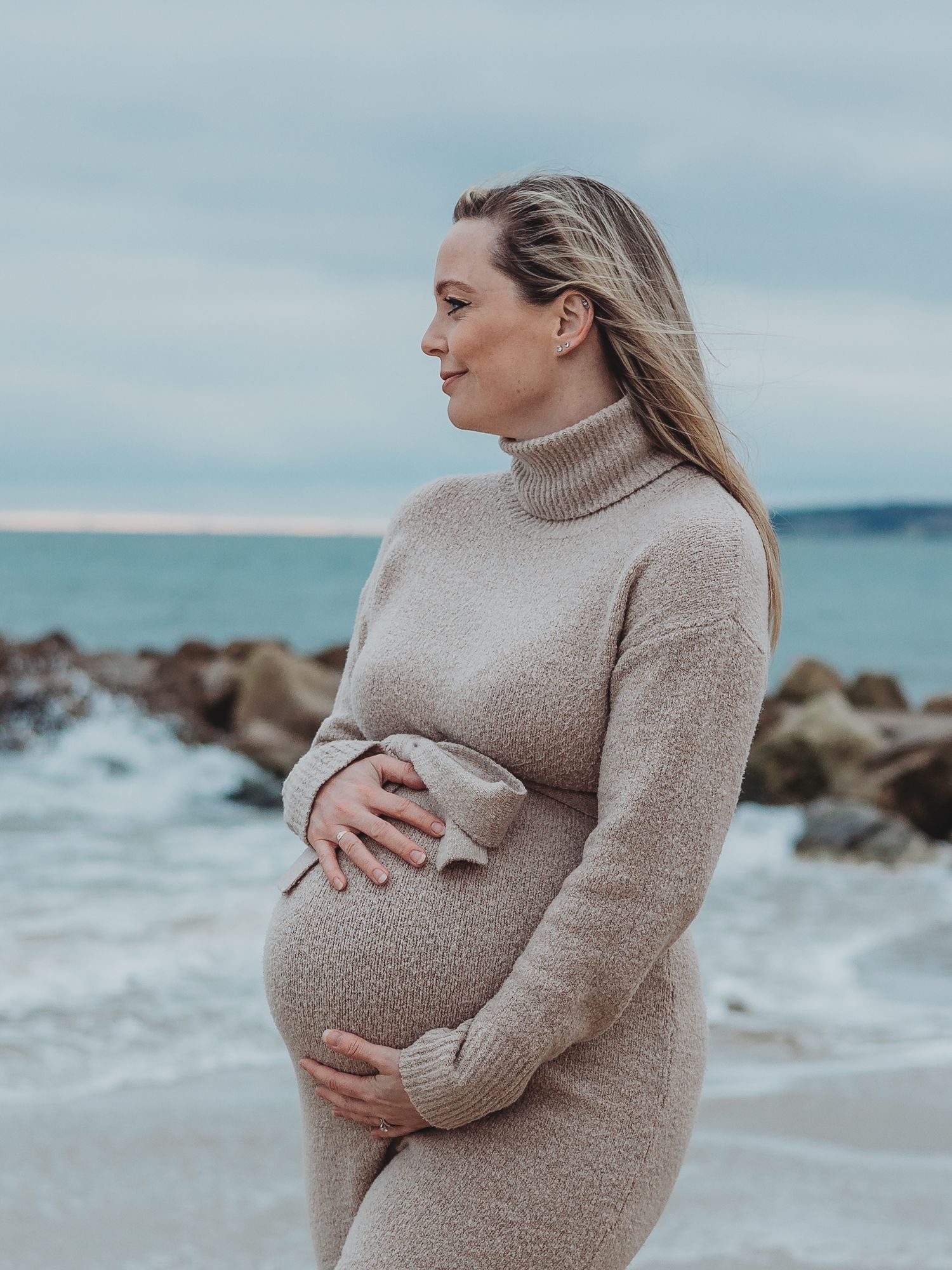 A person in a beige turtleneck sweater stands by the ocean, cradling their pregnant belly against a rocky shoreline.