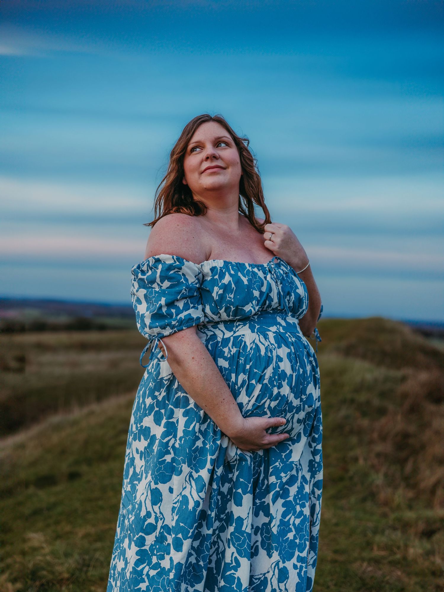 A woman in a blue and white floral dress stands against a scenic outdoor backdrop with rolling hills and blue sky.