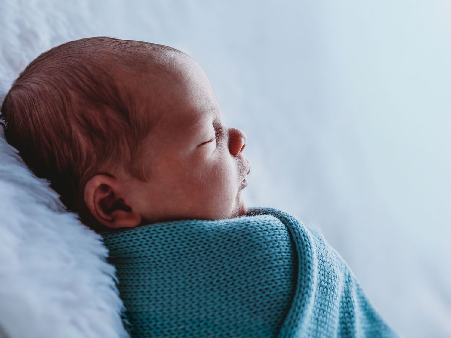 A sleeping newborn baby wrapped in a teal colored swaddle blanket against a white background.