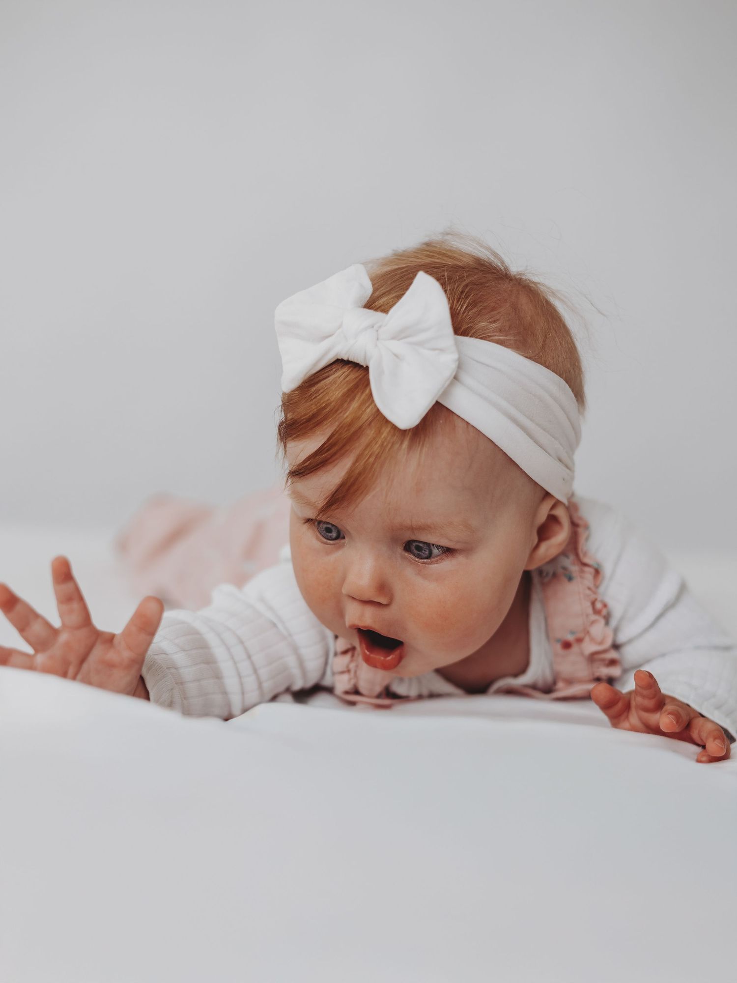 A series of four connected photos shows a baby in white clothing and bow headband laying on light colored bedding.