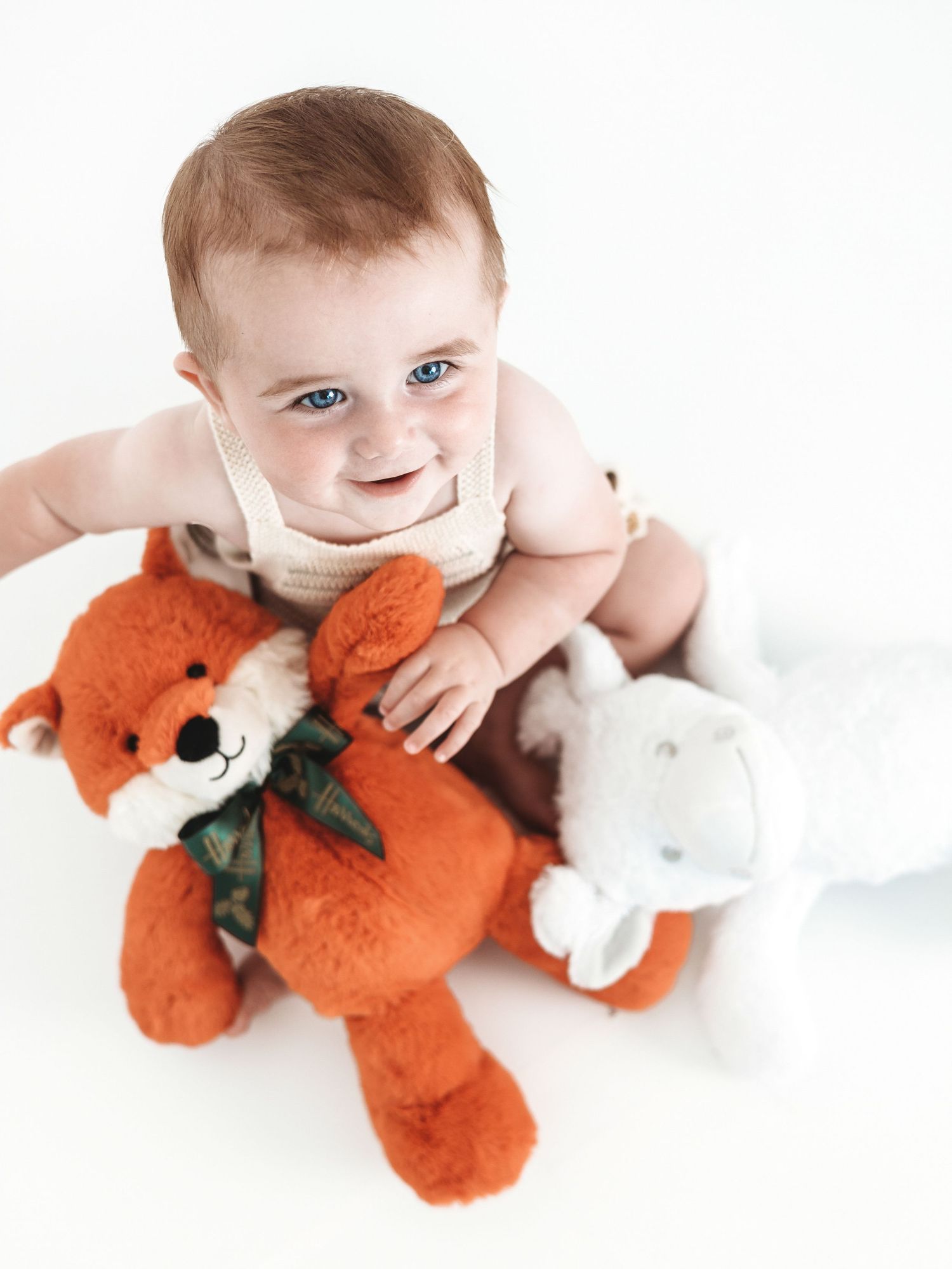 A baby in white clothing cuddles with orange fox and white bunny plush toys on a light background.