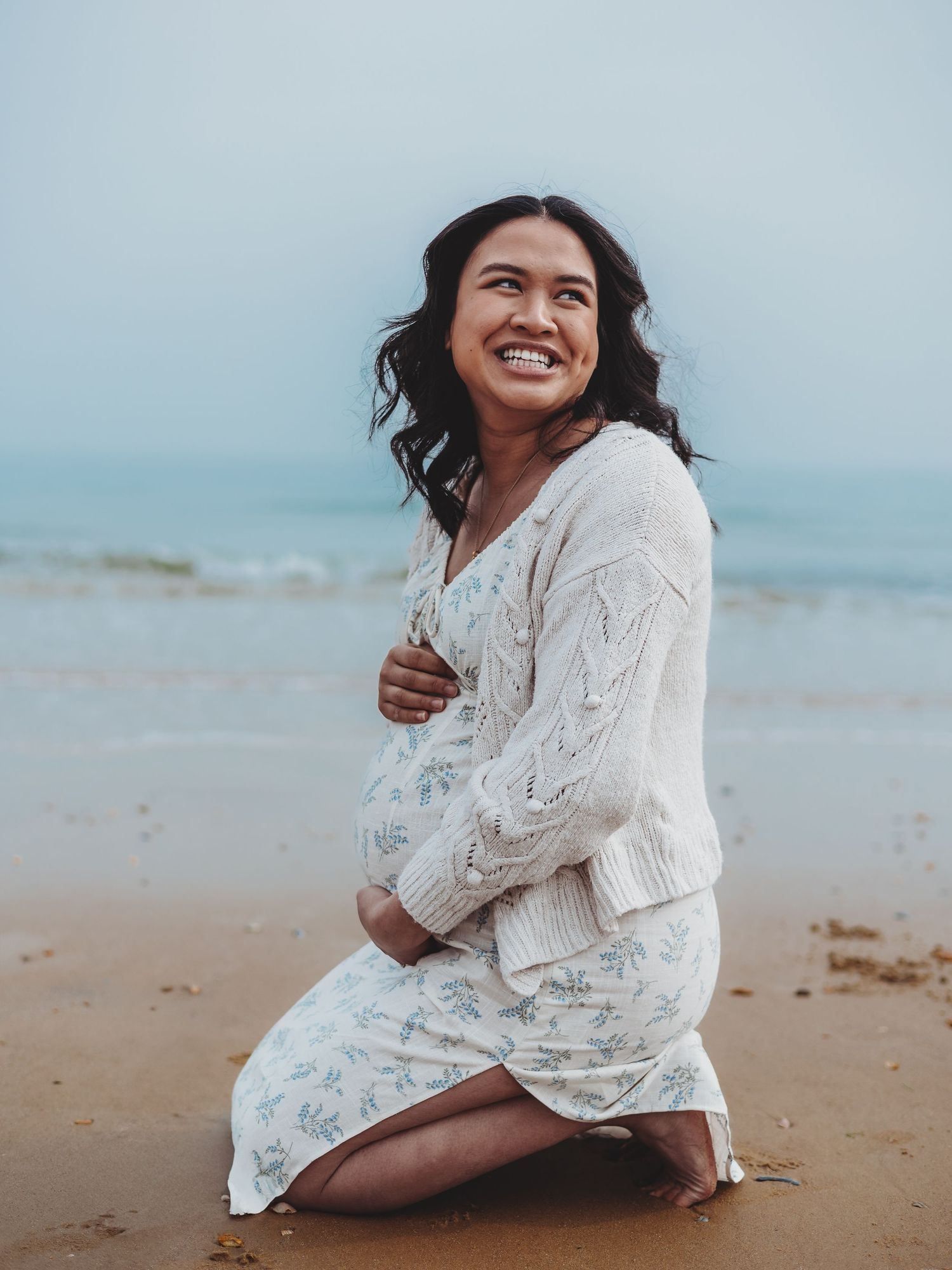 Person in white lace dress sitting on sandy beach at sunset with ocean waves in background.