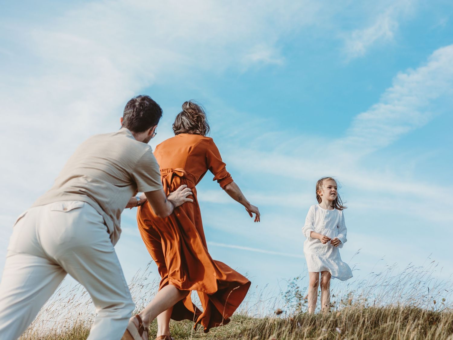 A couple runs playfully through a grassy field on a sunny day with blue skies overhead.