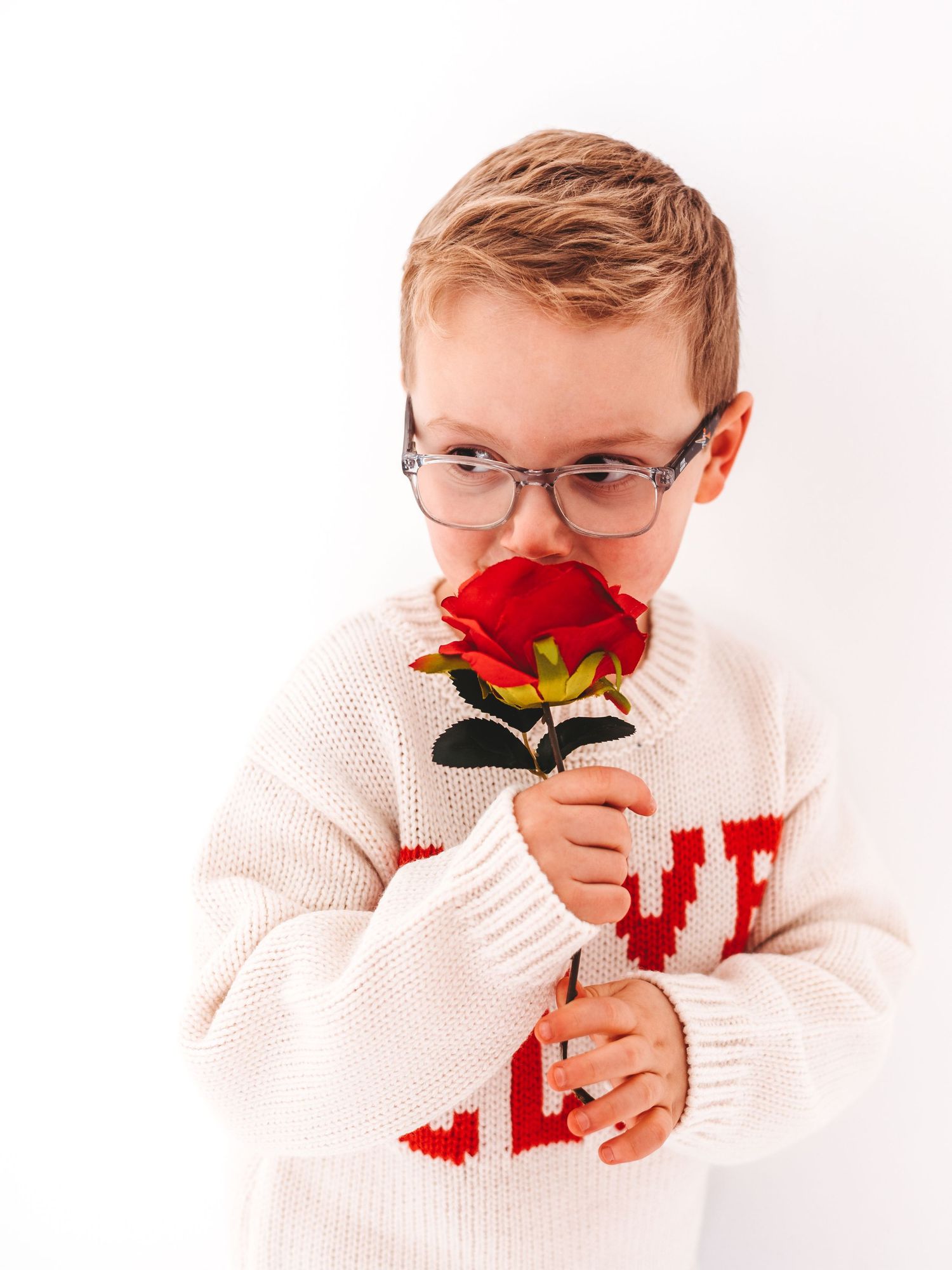 A young boy wearing glasses and a white knit sweater smells a red rose against a white background.