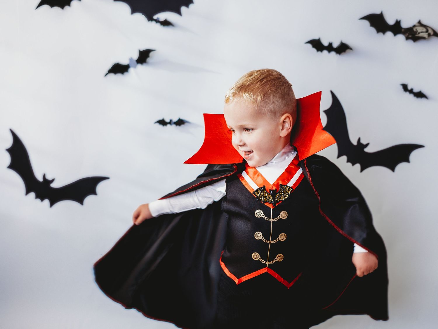 A young boy dressed in a Dracula vampire Halloween costume with black cape and red collar poses with paper bats.