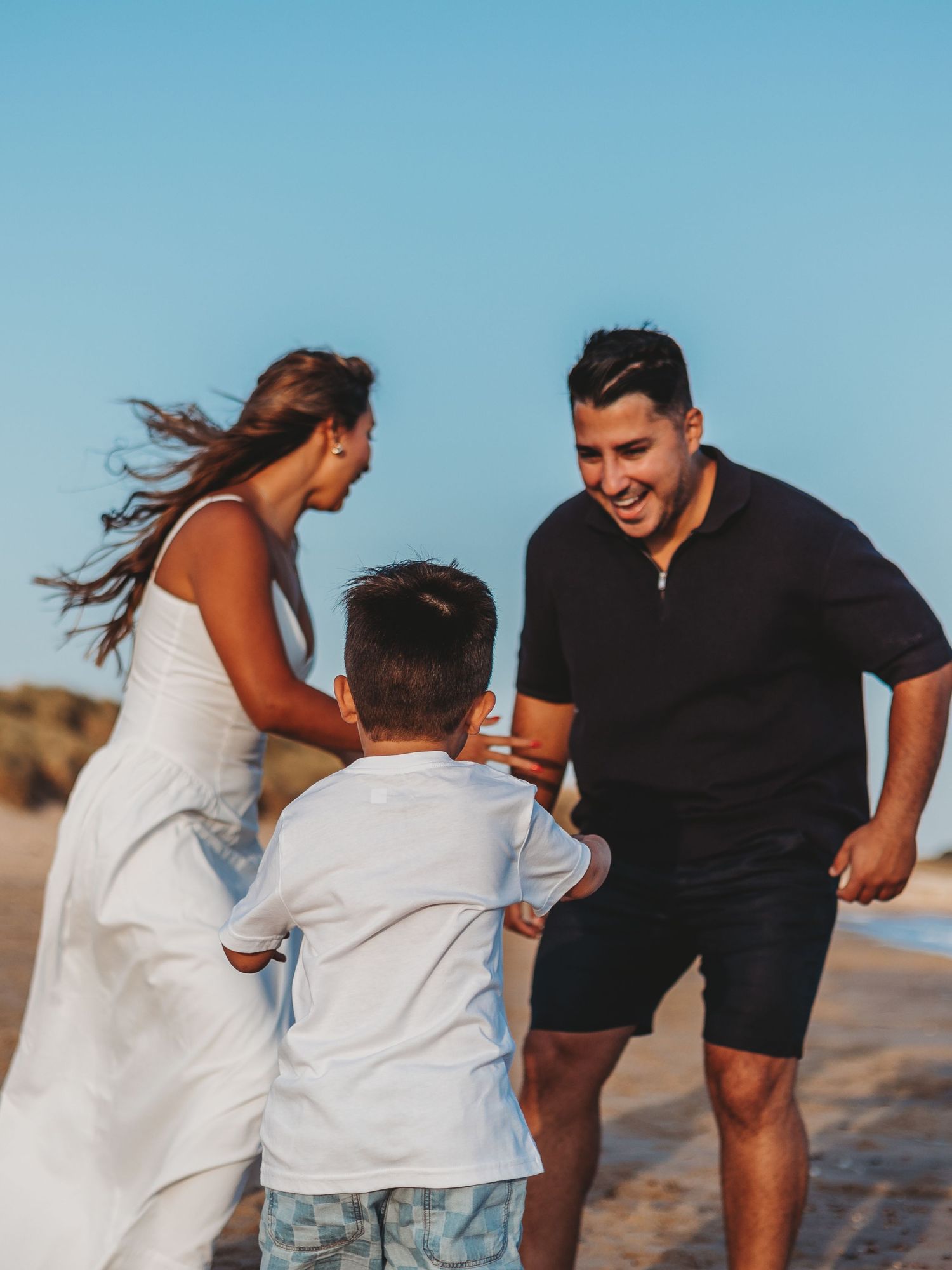 Family of three playing joyfully on a sunny beach, parents smiling at young boy running toward them.