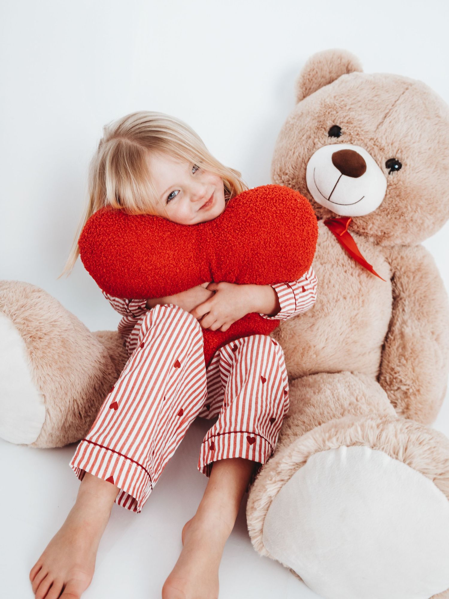 Young girl in striped pajamas hugging a red heart pillow beside a large teddy bear on white background.