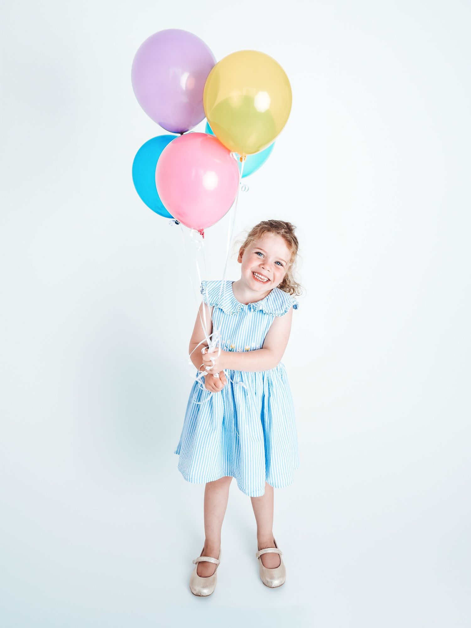 Young girl in light blue dress smiling and holding colorful balloons on white background.
