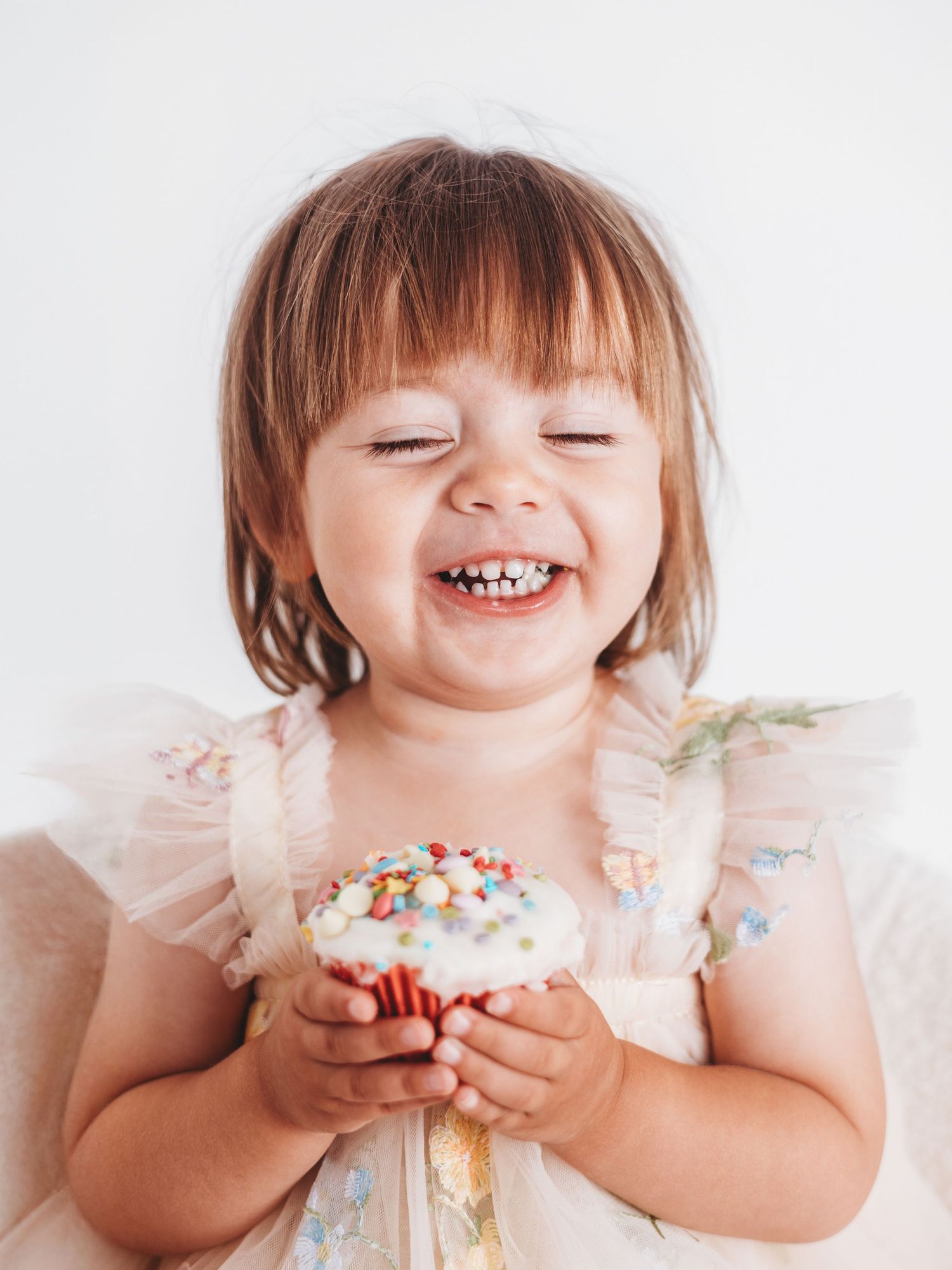 A joyful toddler girl laughing while holding a white frosted cupcake with colorful sprinkles, wearing a pink dress.