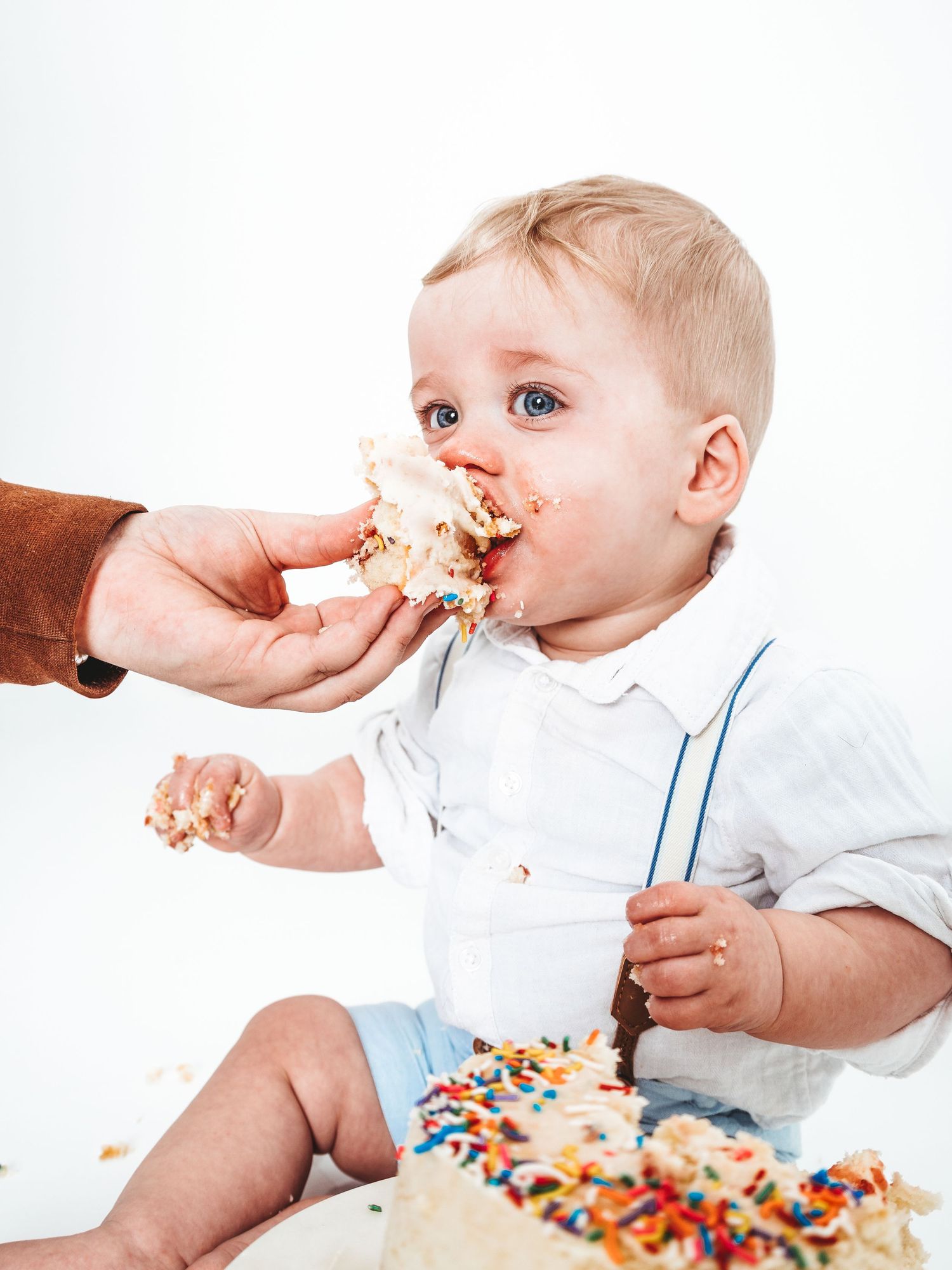 Messy baby boy eating birthday cake smash with sprinkles, blue eyes, white shirt.