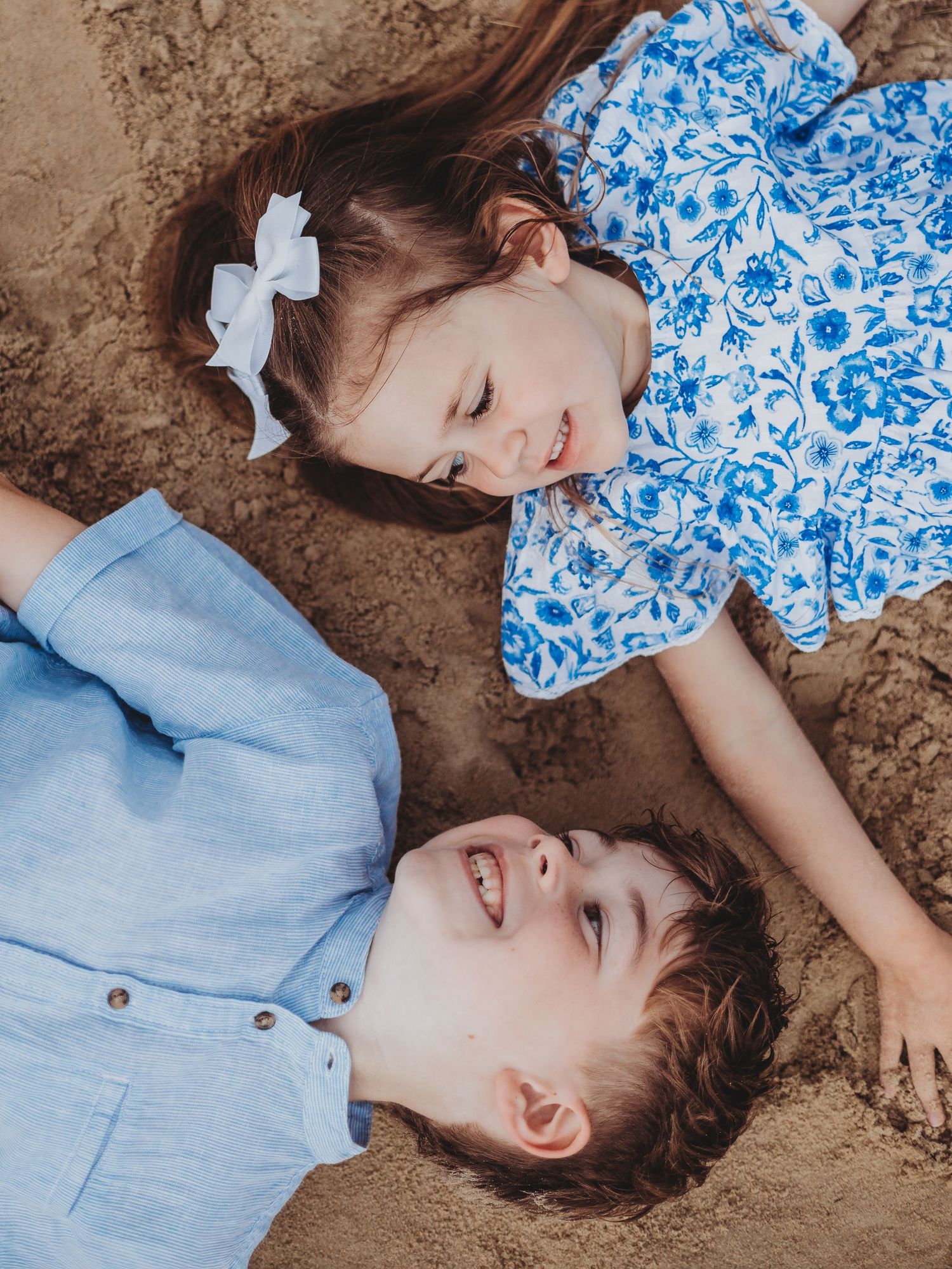 Two young children lying on sand, smiling, wearing coordinating blue outfits, girl with white bow in hair.