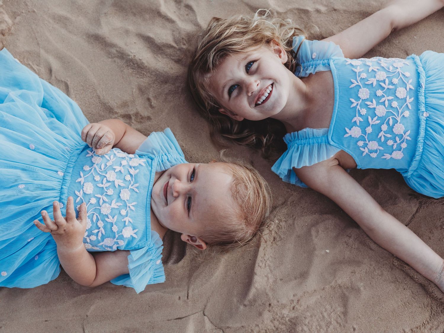 Two young girls in matching blue floral dresses lying on sandy beach, smiling joyfully in coordinating outfits.