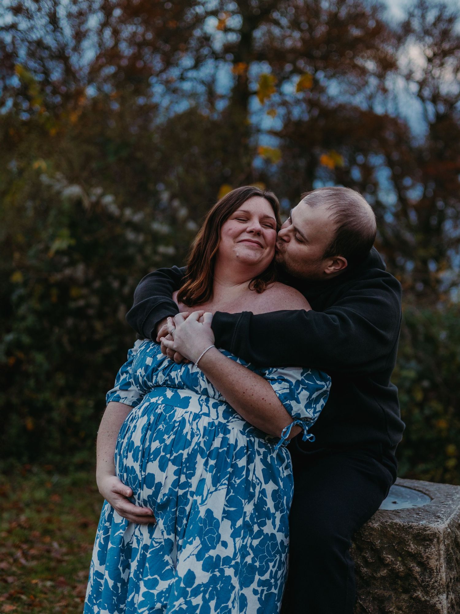 A couple in a blue floral dress and black jacket embrace lovingly against an autumn forest backdrop.