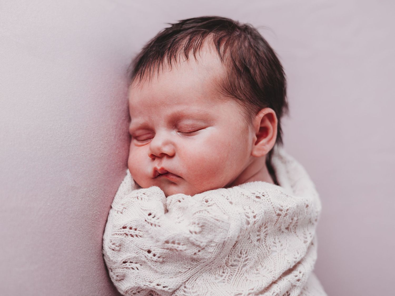 A sleeping newborn baby wrapped in white lace swaddle blanket rests peacefully against a light background.