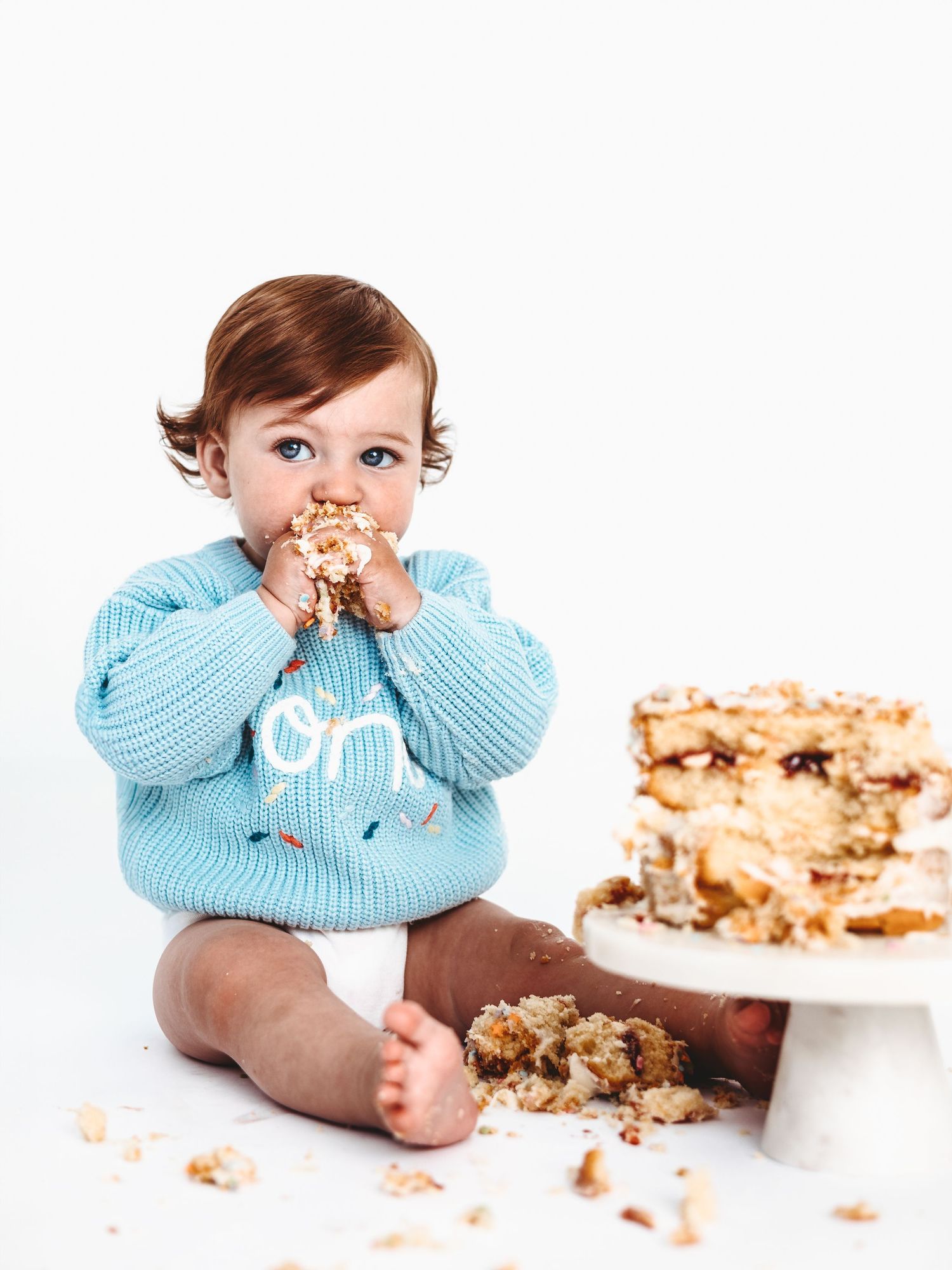 Baby in light blue sweater sits with birthday cake and enjoys messy first birthday cake smash photo session.