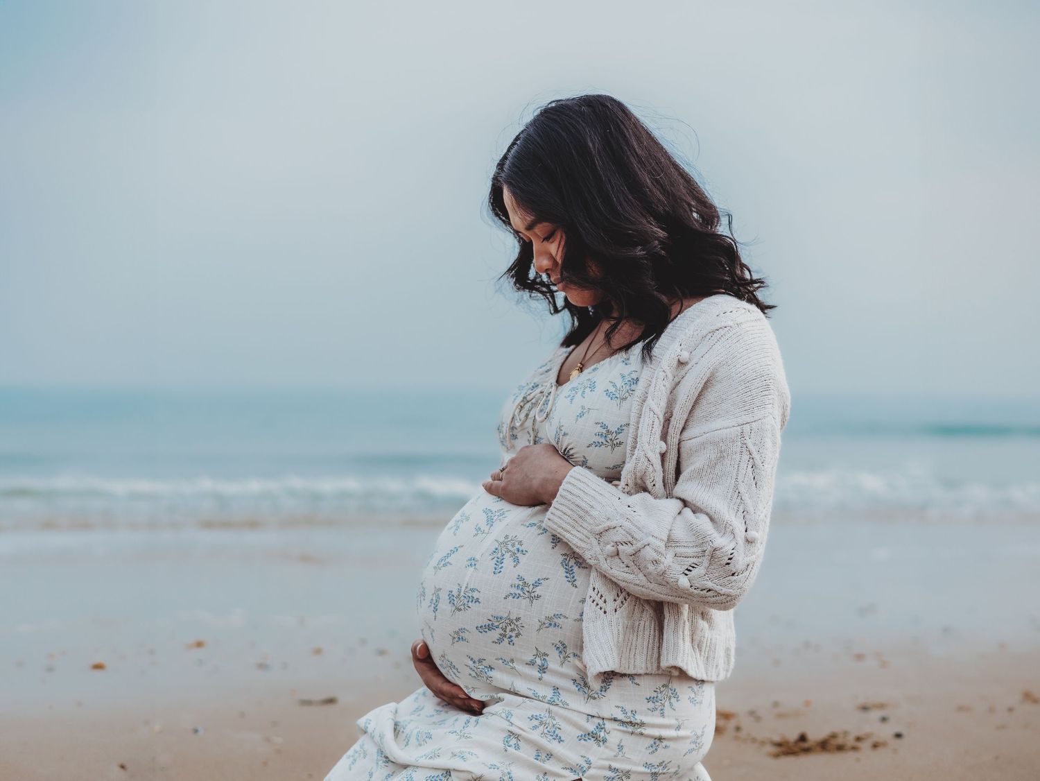Pregnant woman in white dress stands on beach at sunset holding her belly in series of maternity photos.