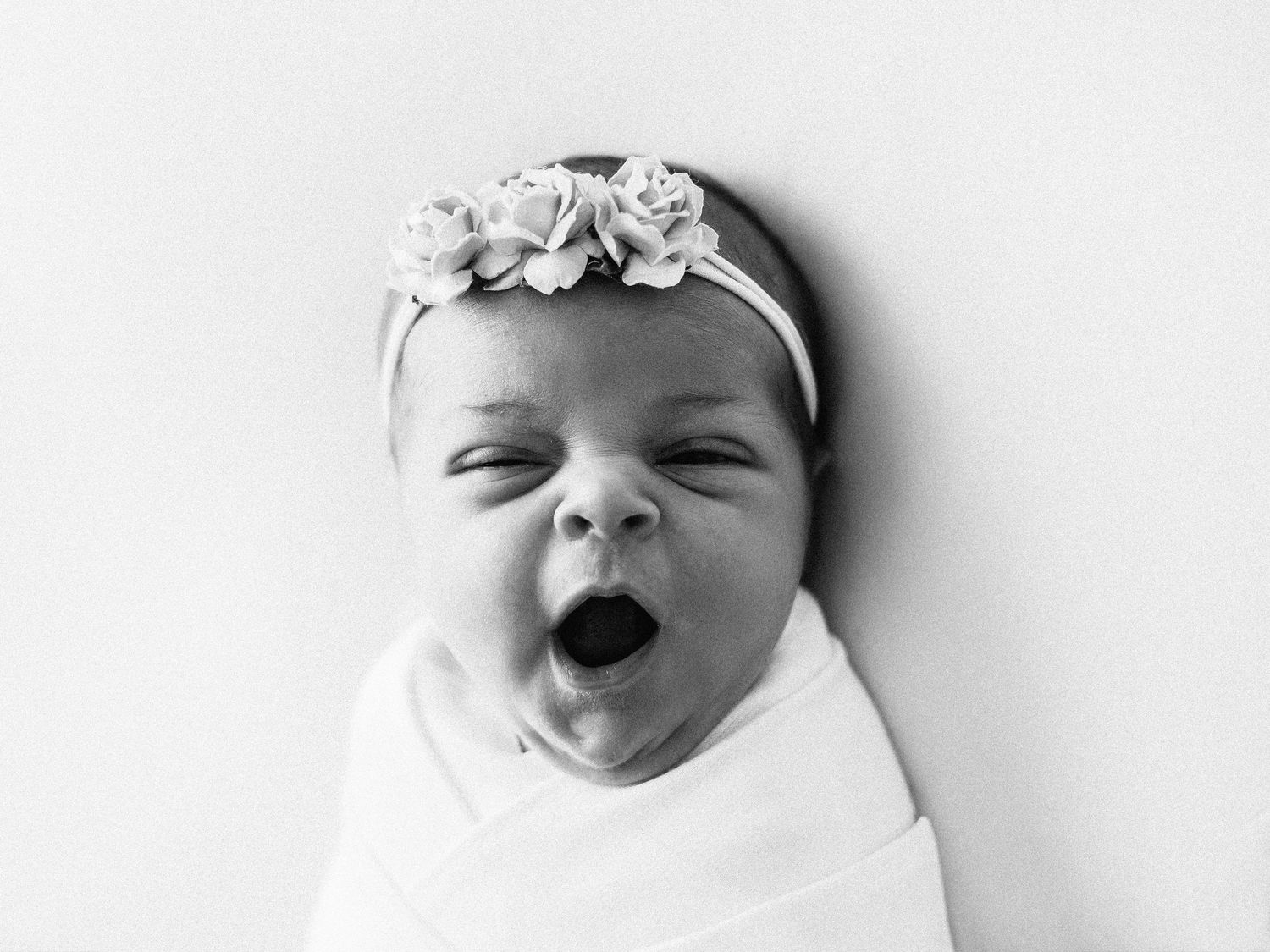 A black and white photo of a yawning newborn baby wearing a floral headband and swaddled in white fabric.