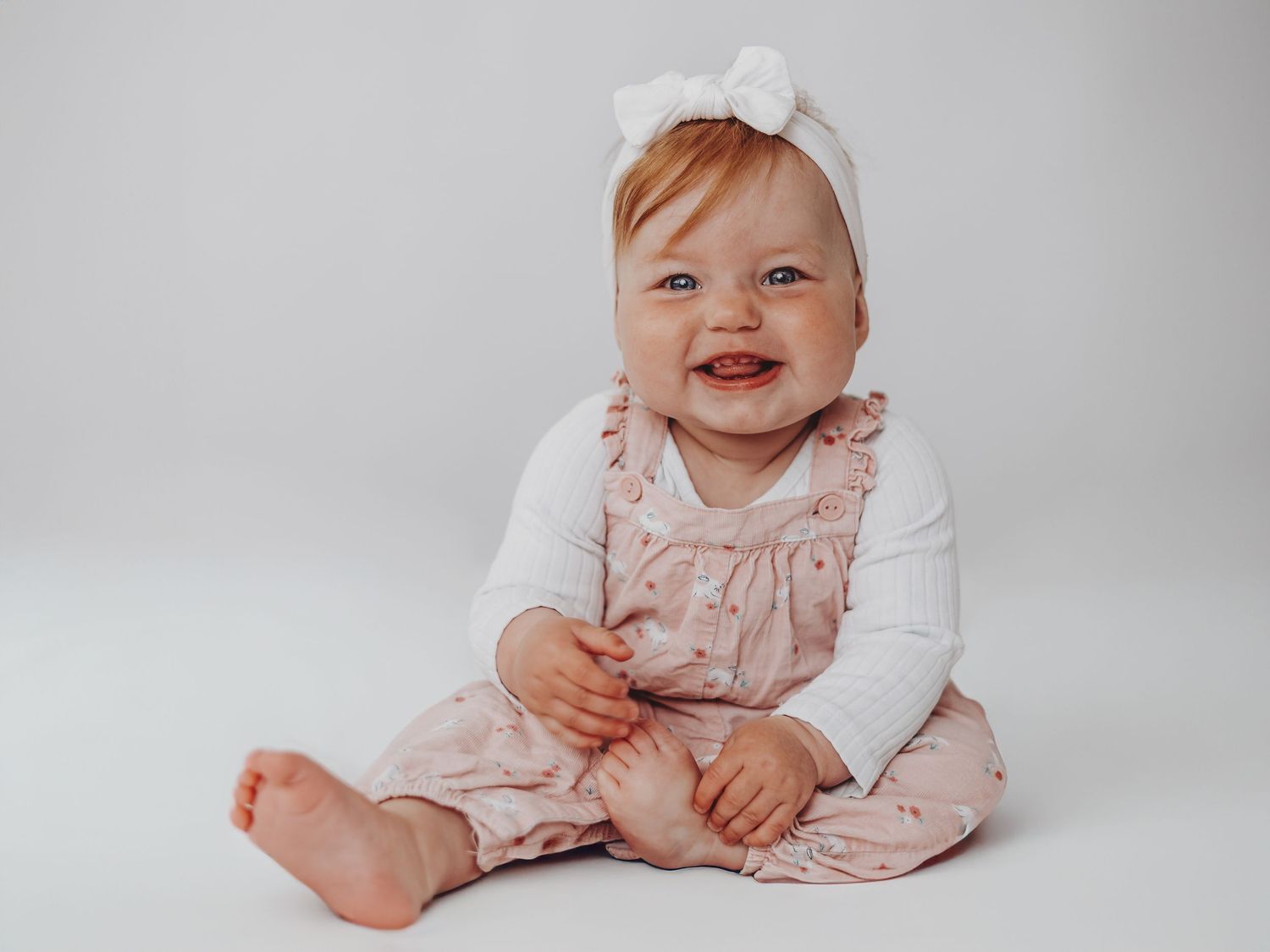 A happy baby wearing a white bow, white long sleeve shirt and pink overalls sits against a light gray background.