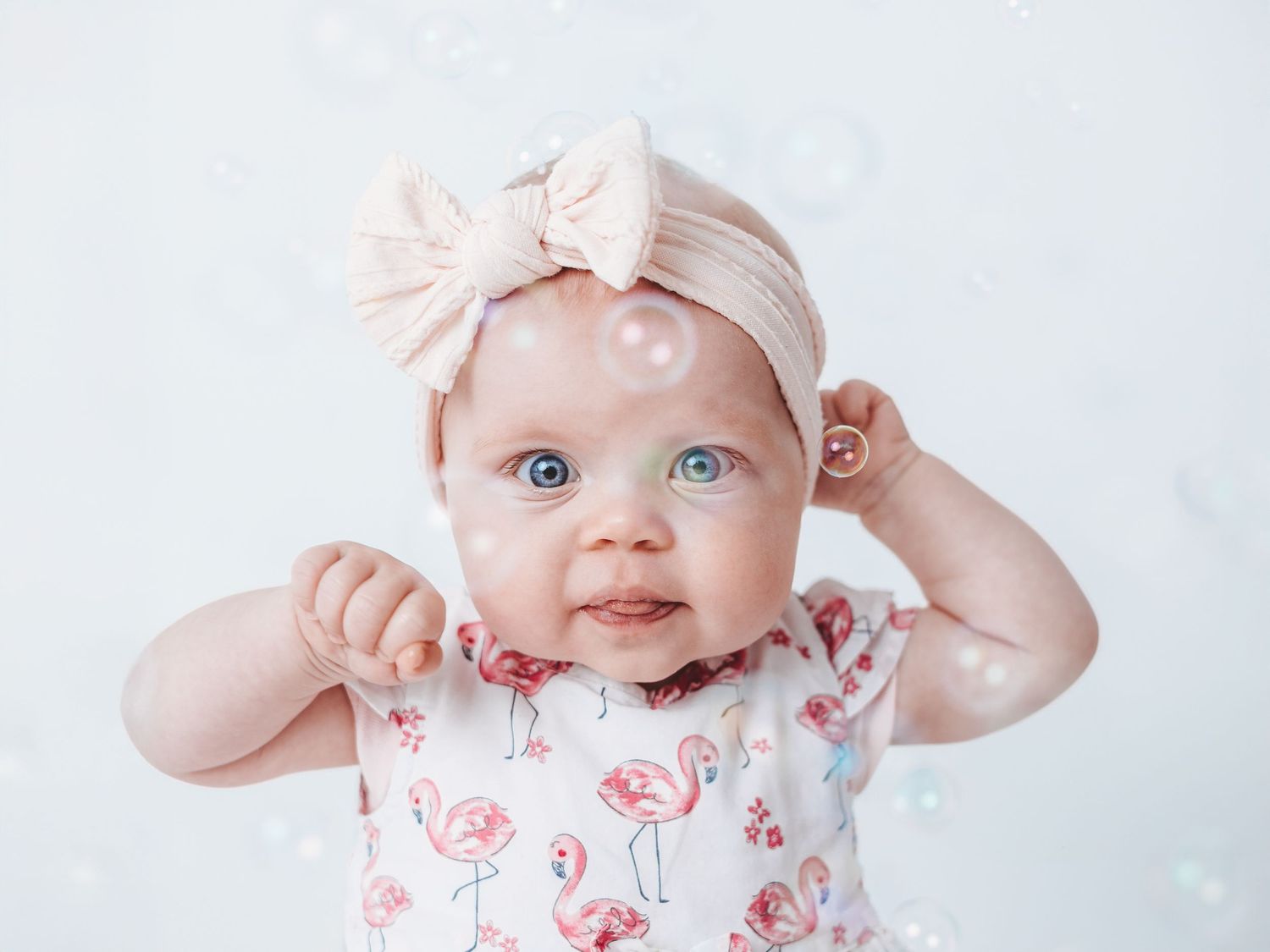 Baby wearing floral outfit and white bow headband lies on white blanket while smiling at camera.