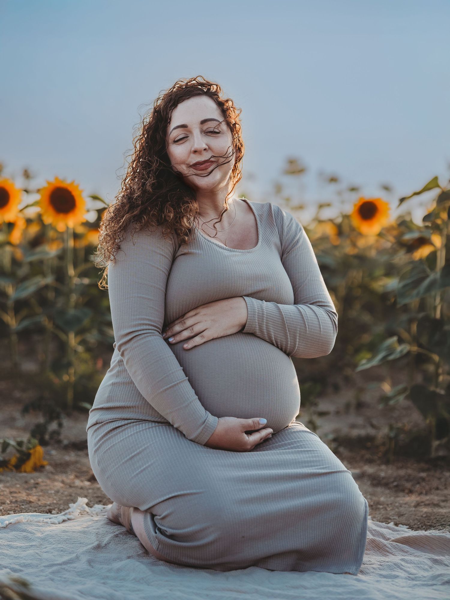 Expectant mother in a gray dress sits peacefully among blooming sunflowers during golden hour maternity photoshoot.