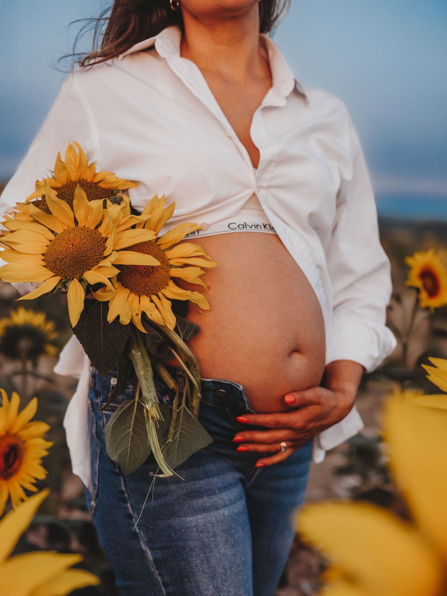 Person in white button-down shirt and jeans poses in sunflower field during golden hour.