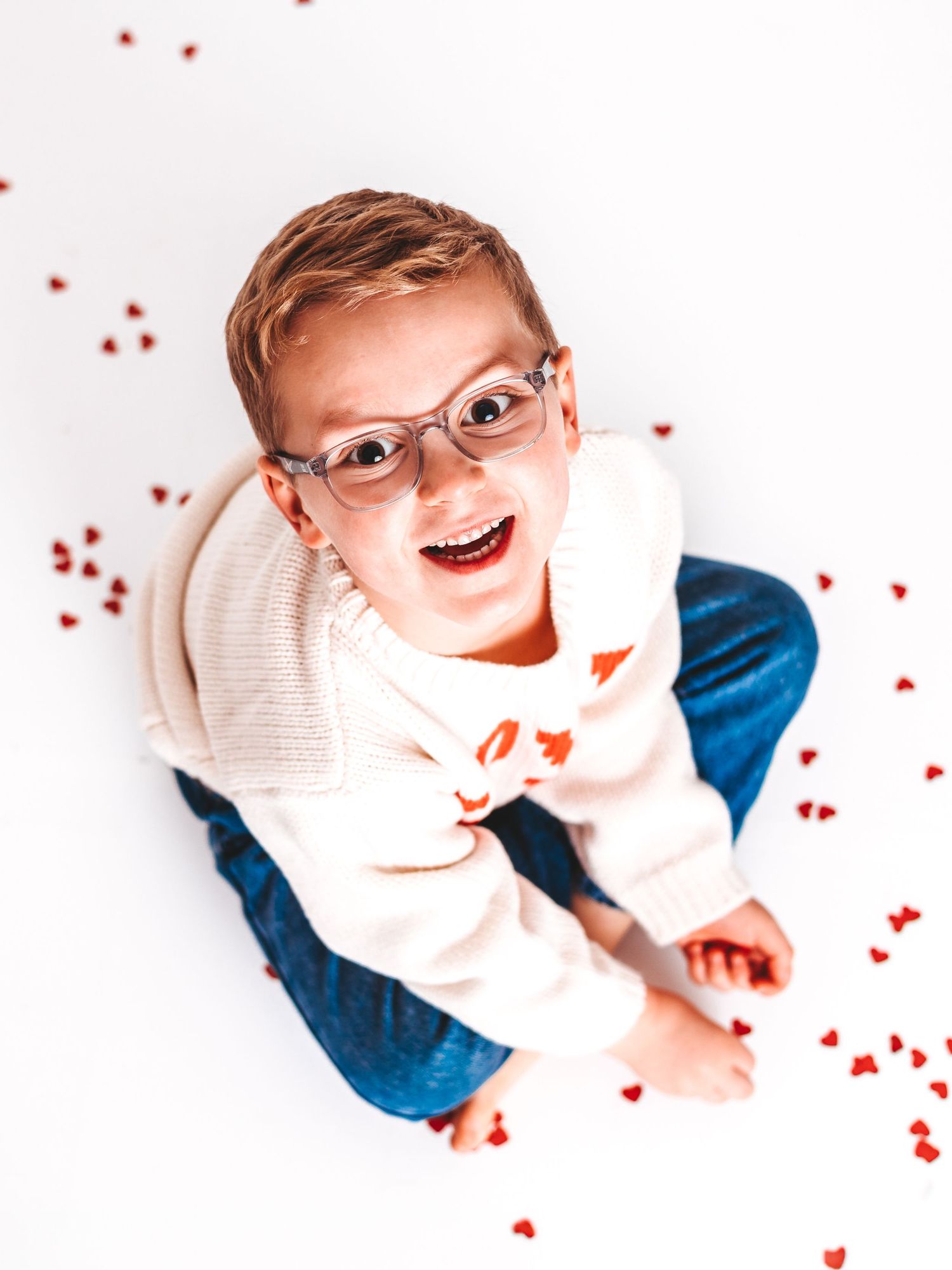A joyful child with glasses sits on a white floor surrounded by red heart confetti, wearing a white sweater and blue jeans.