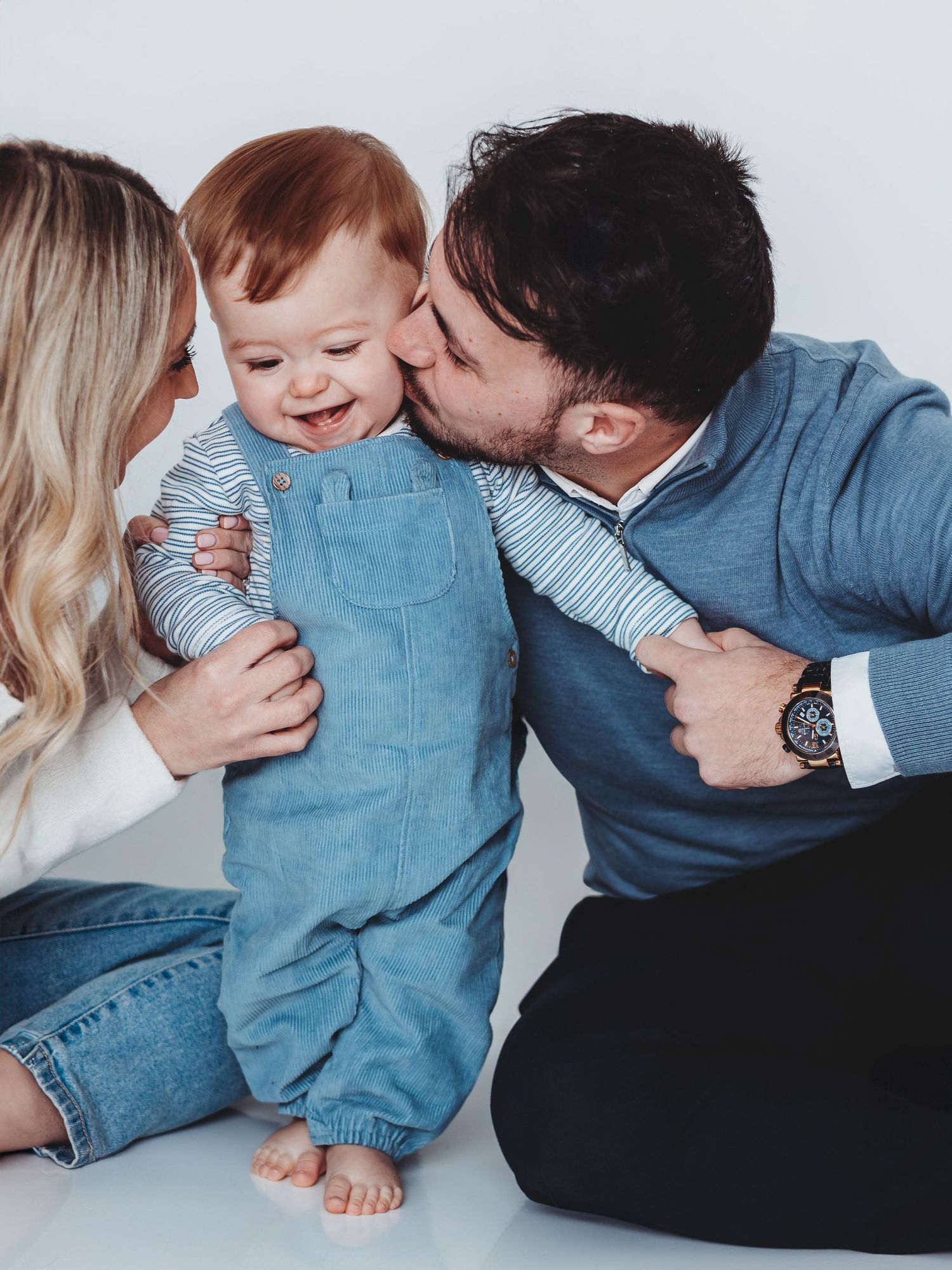 Happy baby laughing as parents kiss cheeks in a sweet family portrait studio photo.