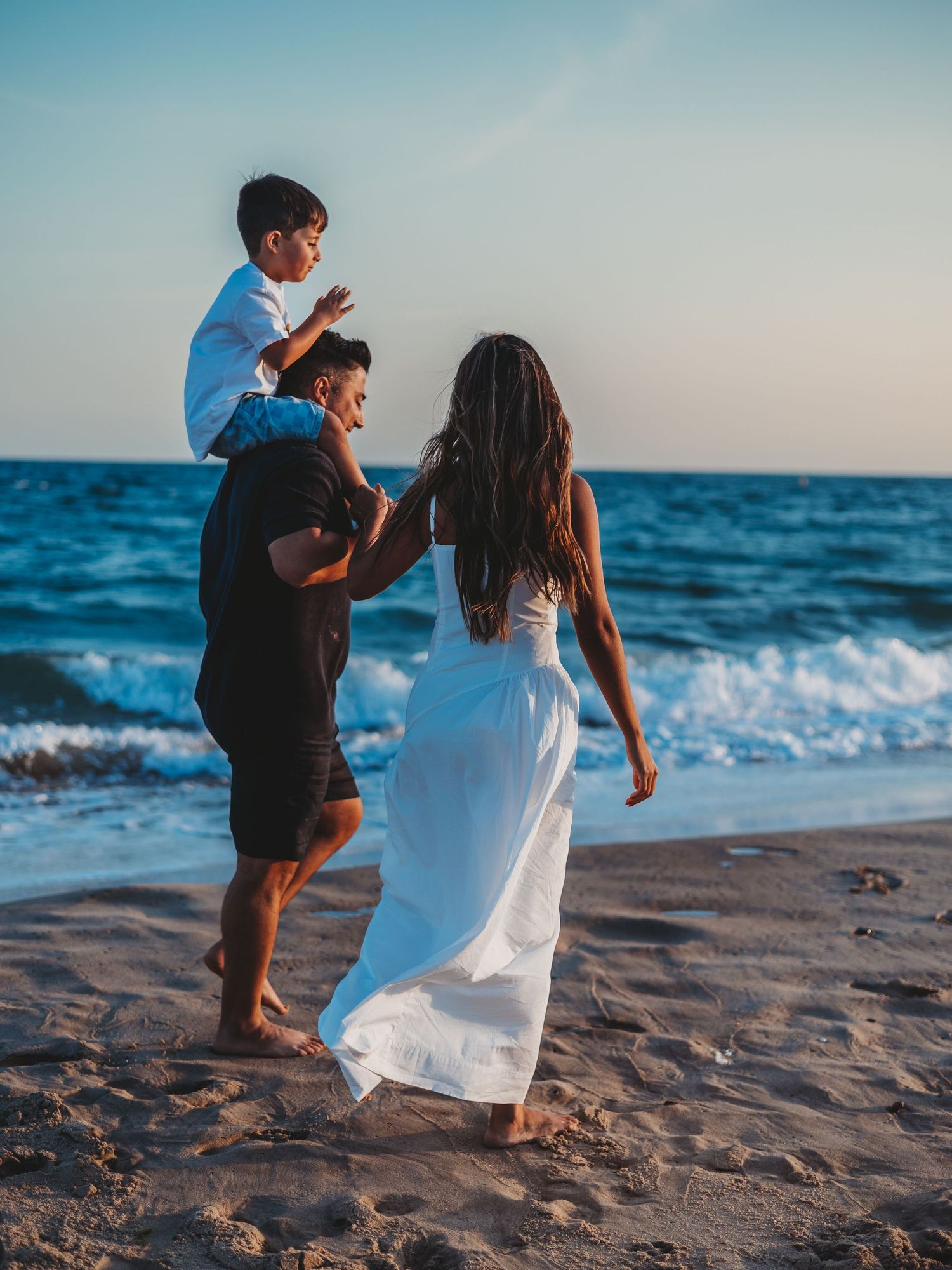 Family enjoying a beach sunset walk, mother in white dress, father carrying son on shoulders, ocean waves in background.