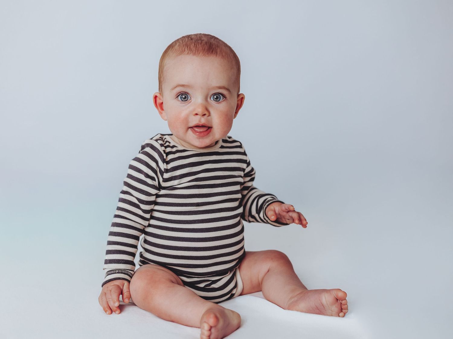 A baby in a black and white striped shirt sits and plays while making joyful expressions.