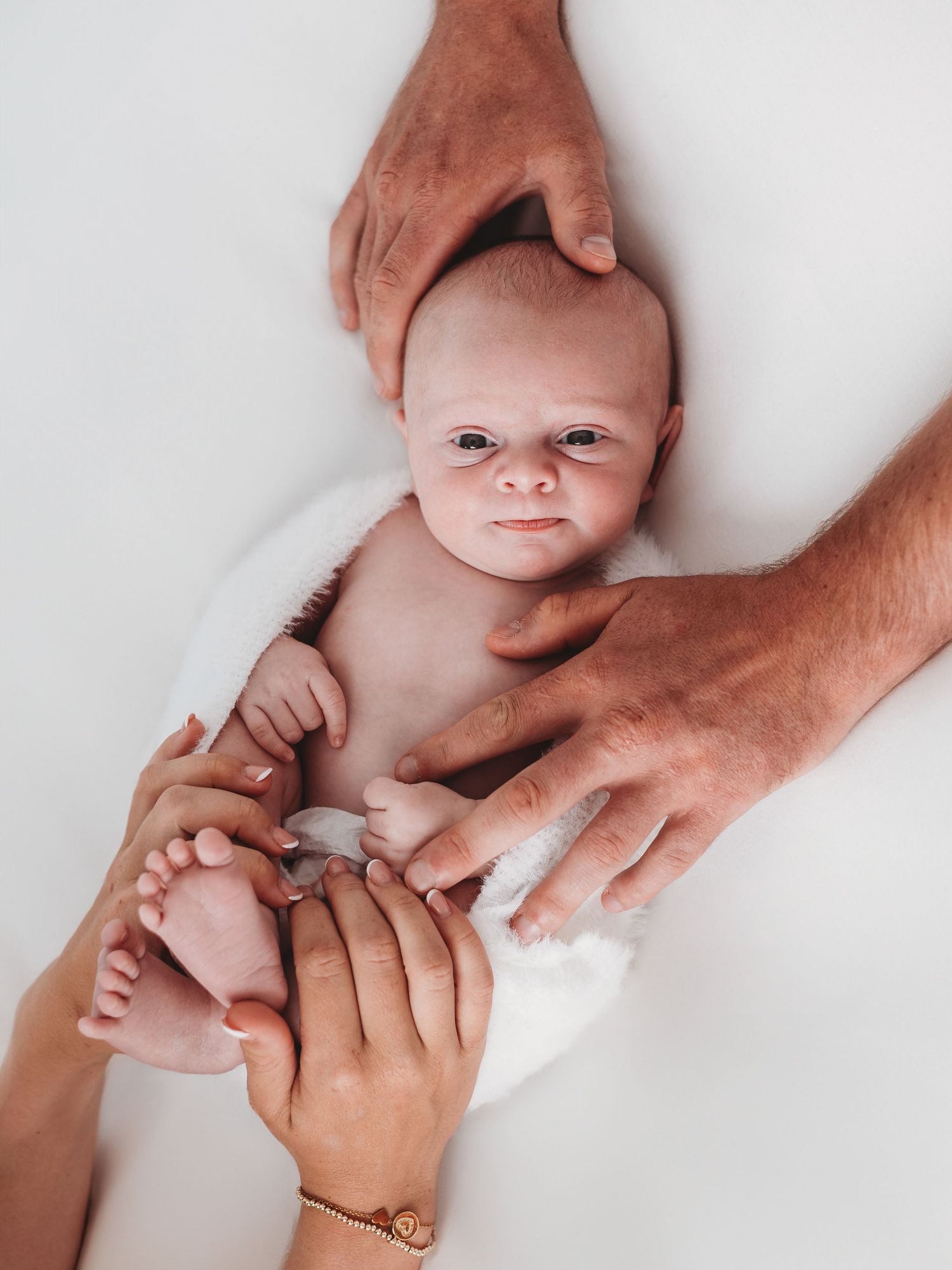 Gentle hands tenderly hold and support a newborn baby lying on a white surface. Dorset Newborn Photoshoot. Poole and Bournemouth.