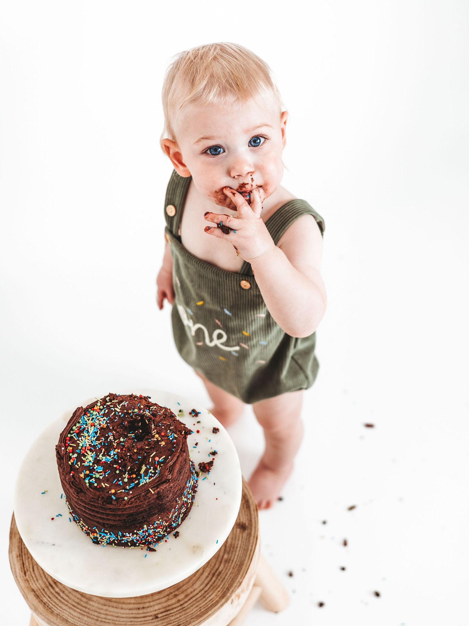 Baby in green romper eating chocolate sprinkle smash cake, celebrating first birthday against white background.