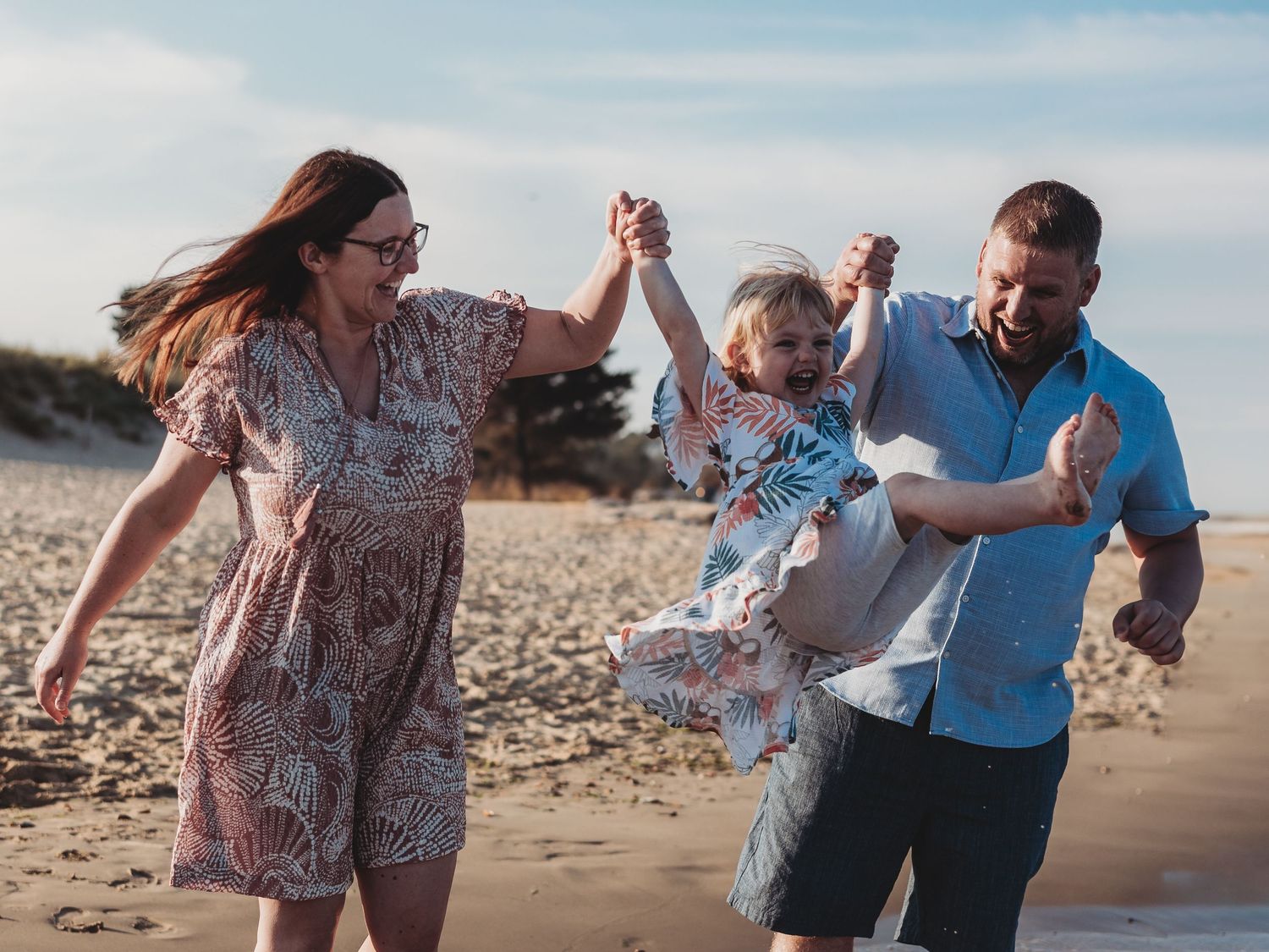 Happy family swinging young child on sunny beach, parents holding hands with toddler, sandy dunes in background.