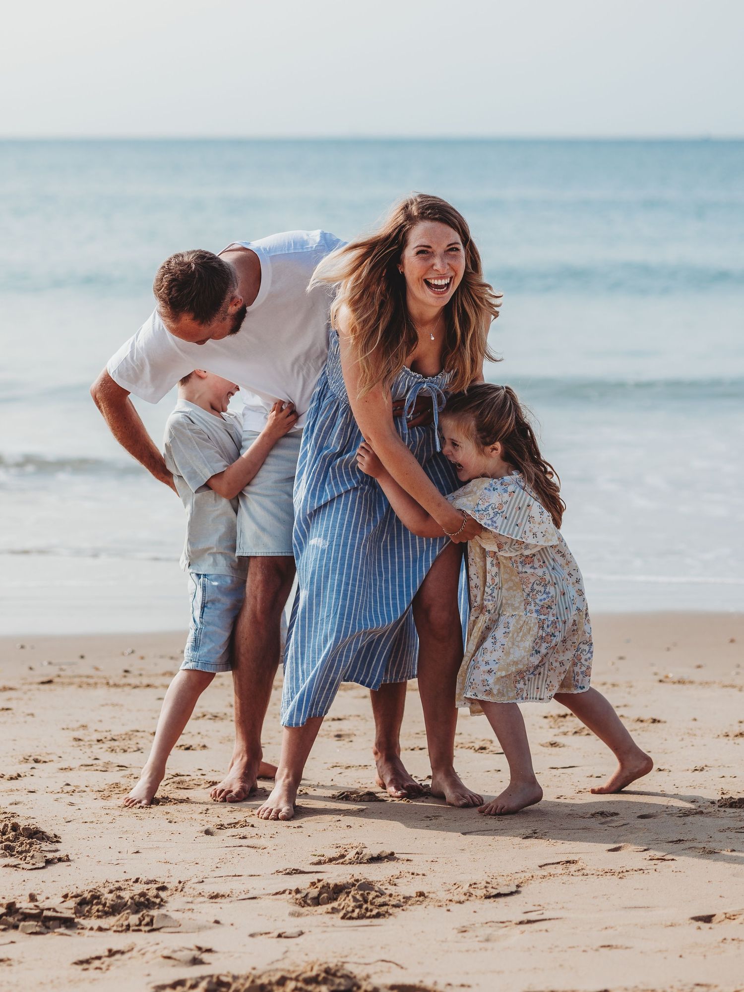 Happy family laughing and playing together on a sandy beach, with two children hugging their smiling parents near ocean waves.