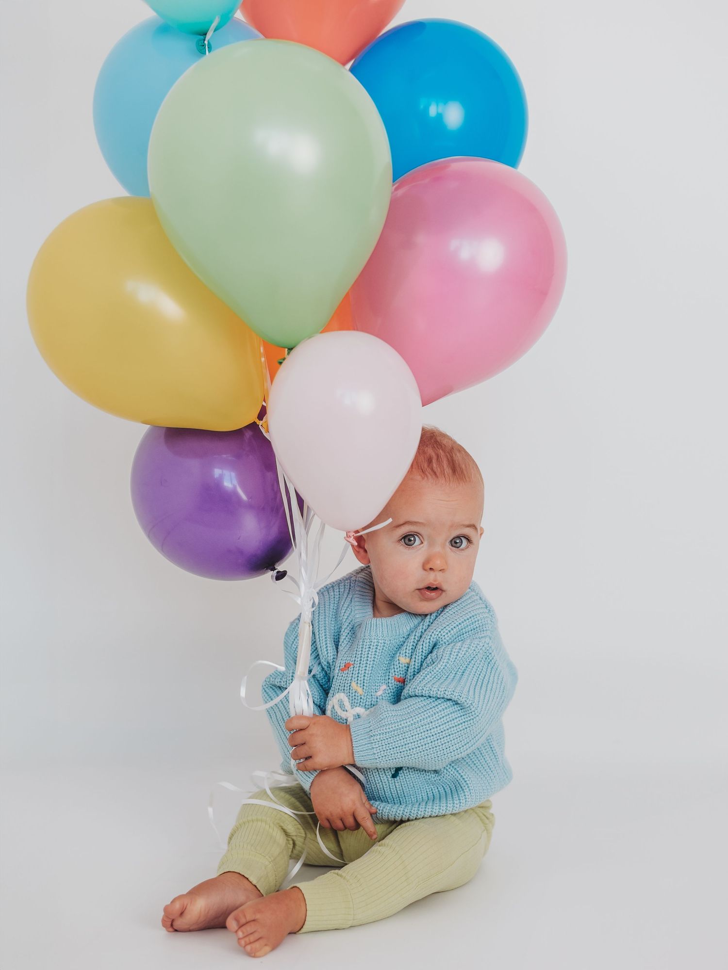 Colorful balloons float beside a small baby wearing a blue sweater and yellow pants against a white backdrop.