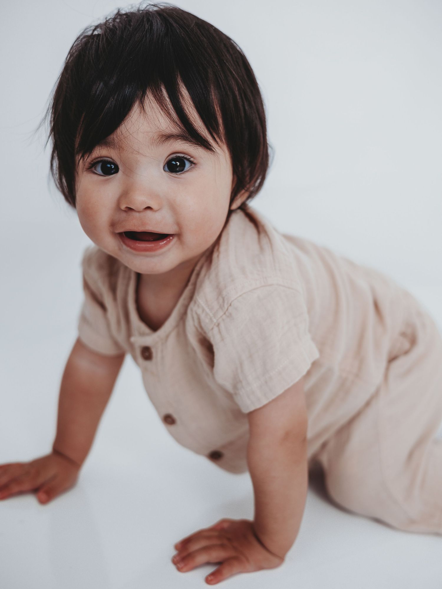 A baby in beige clothing crawls and smiles while being photographed from different angles on a white background.