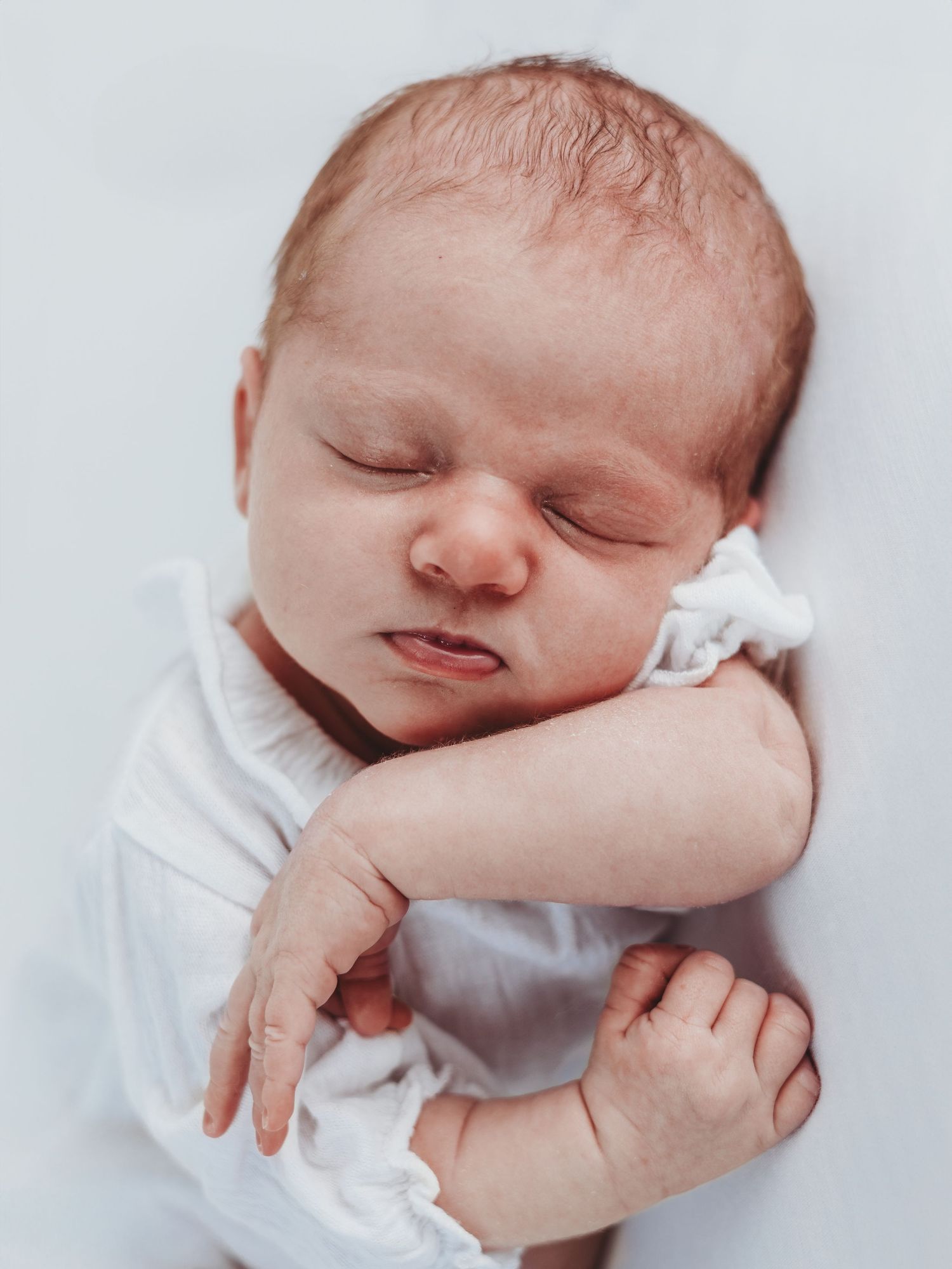 A sleeping newborn baby wearing white is cradled in gentle hands against a light background.