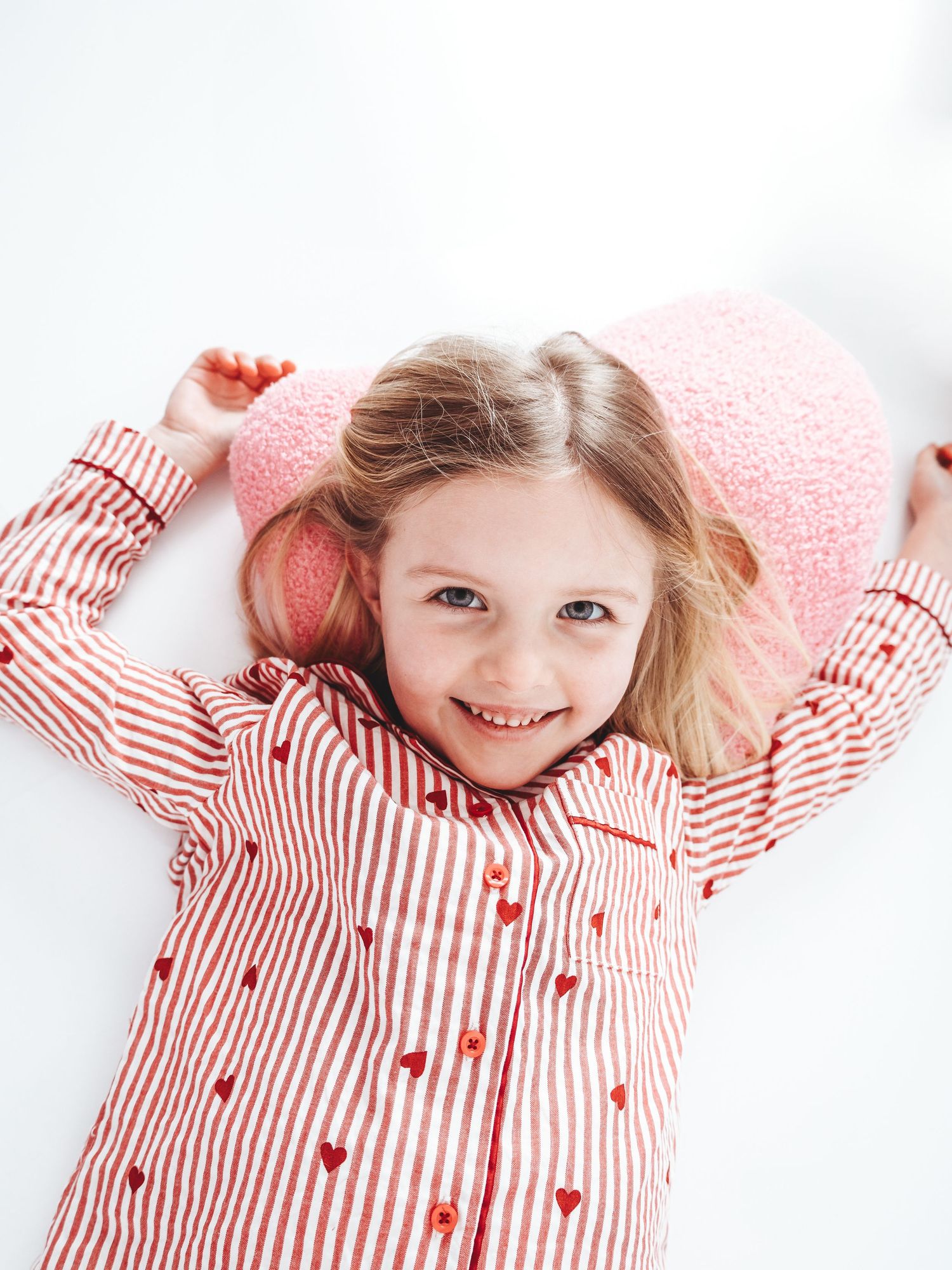 Smiling young girl lying down wearing red striped heart print shirt and pink fuzzy hat with arms raised.
