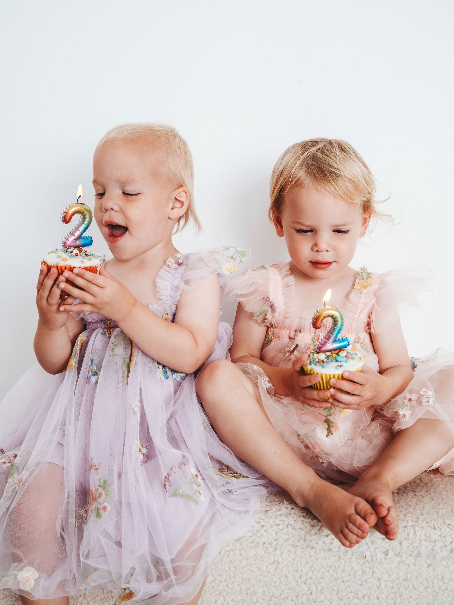 Two toddler twin girls in pink tulle dresses celebrating their 2nd birthday with cupcakes and number 2 candles.