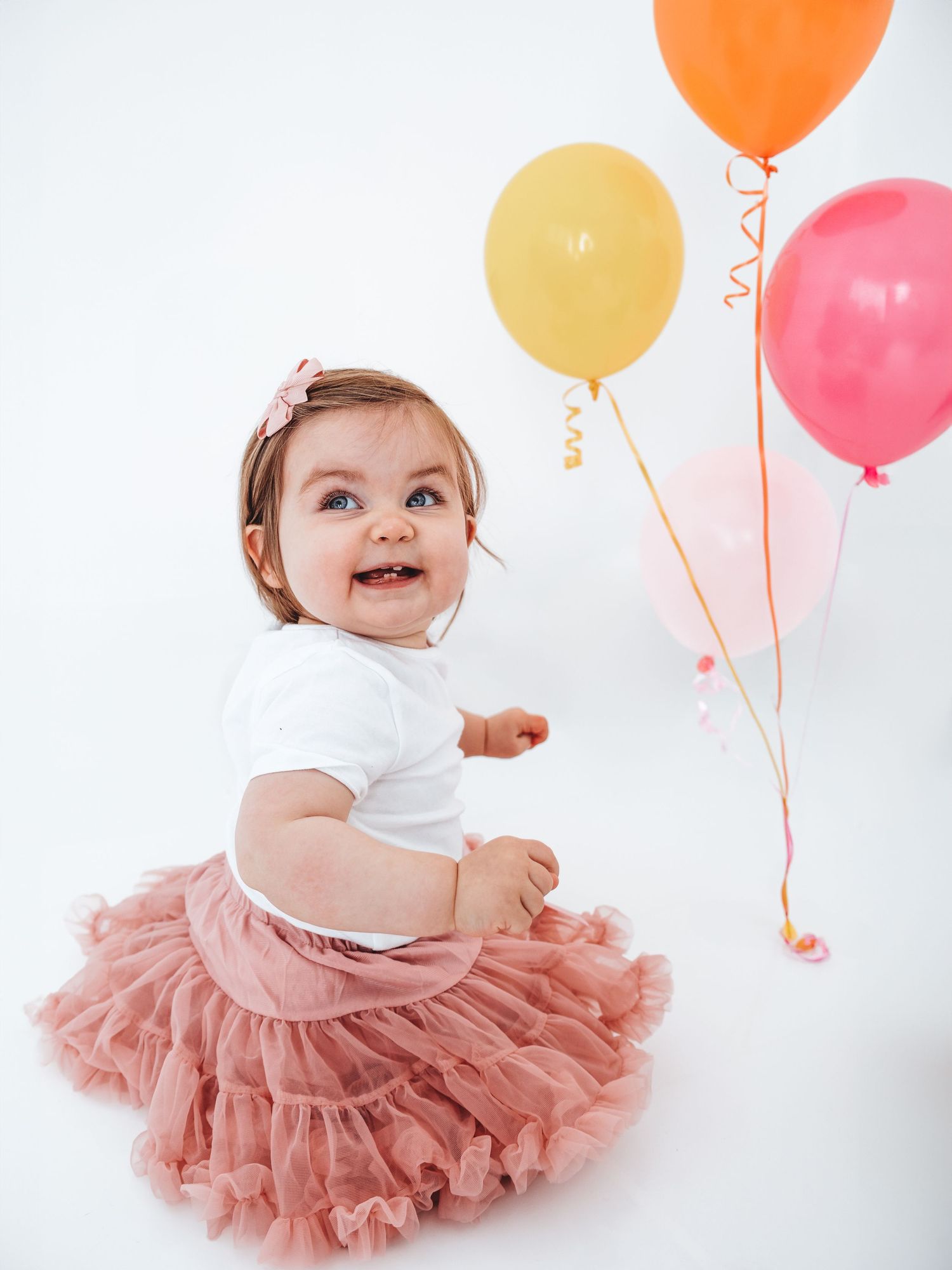 Happy baby girl in pink tutu skirt sitting with colorful balloons at birthday party.
