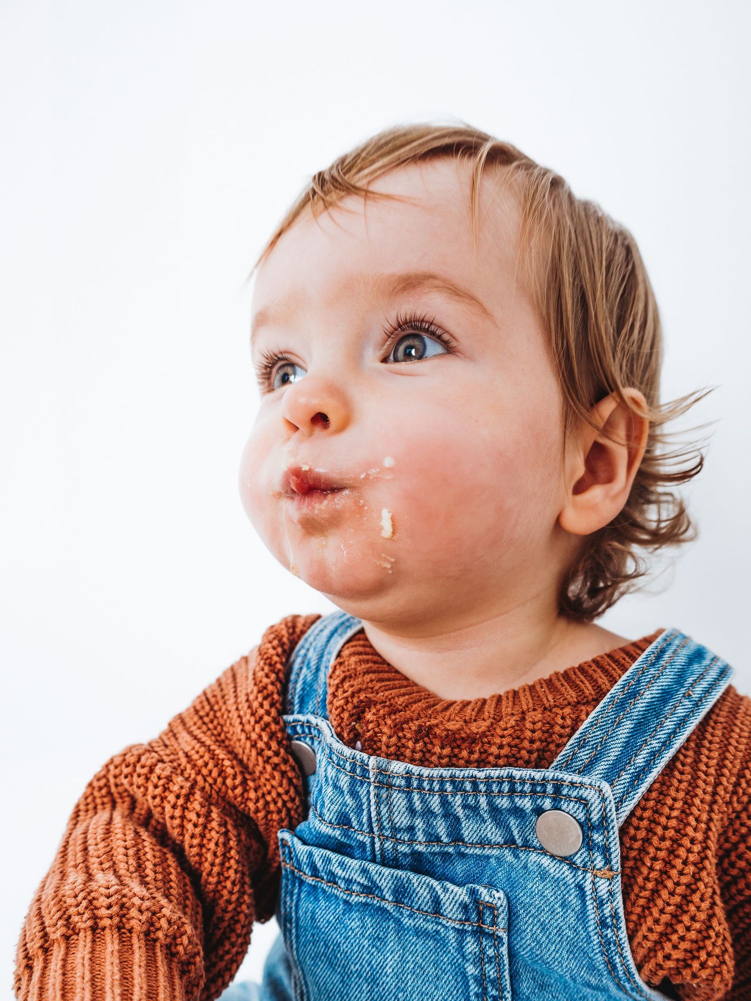 Adorable toddler in denim overalls and rust sweater with food on chin, looking up curiously against white background.
