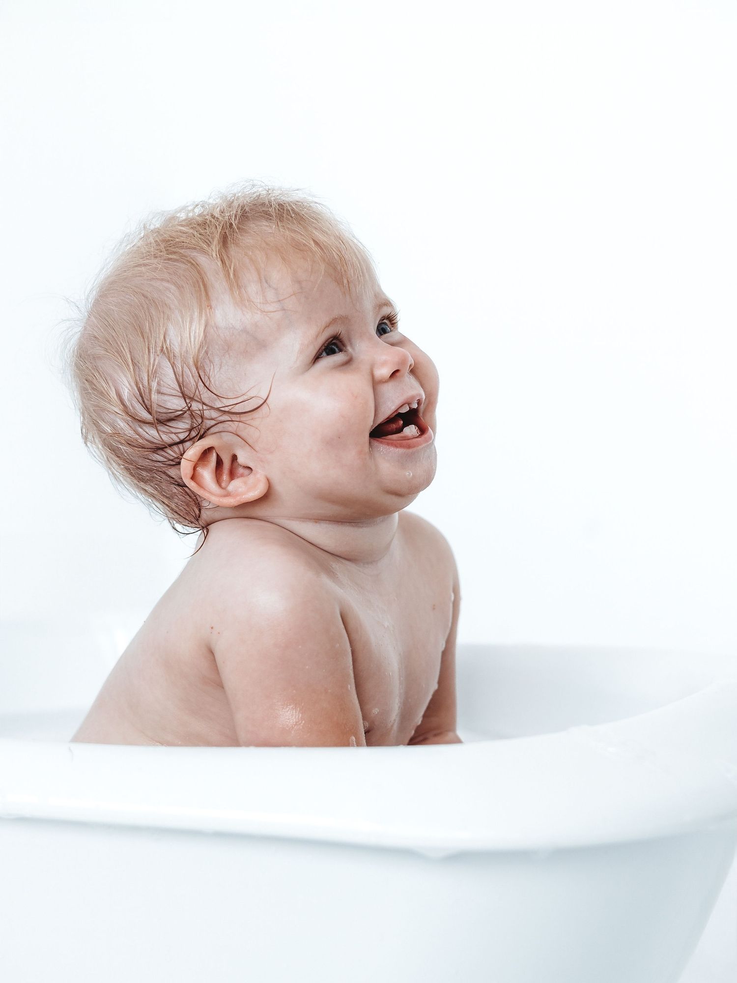 Happy baby with wet hair laughing in a white bathtub during bath time.