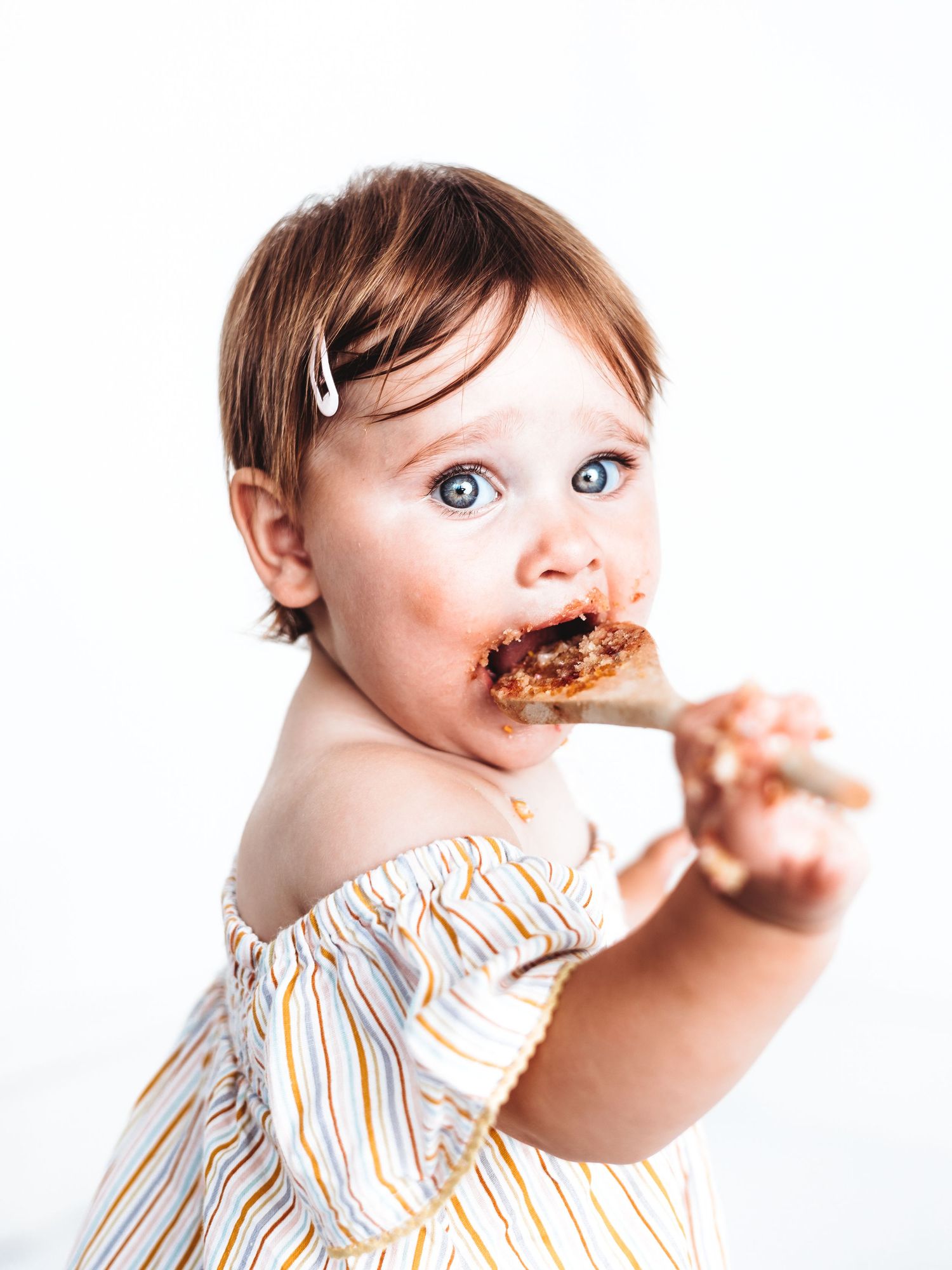 Cute baby girl with blue eyes eating chocolate off a wooden spoon, wearing a striped off-shoulder top.
