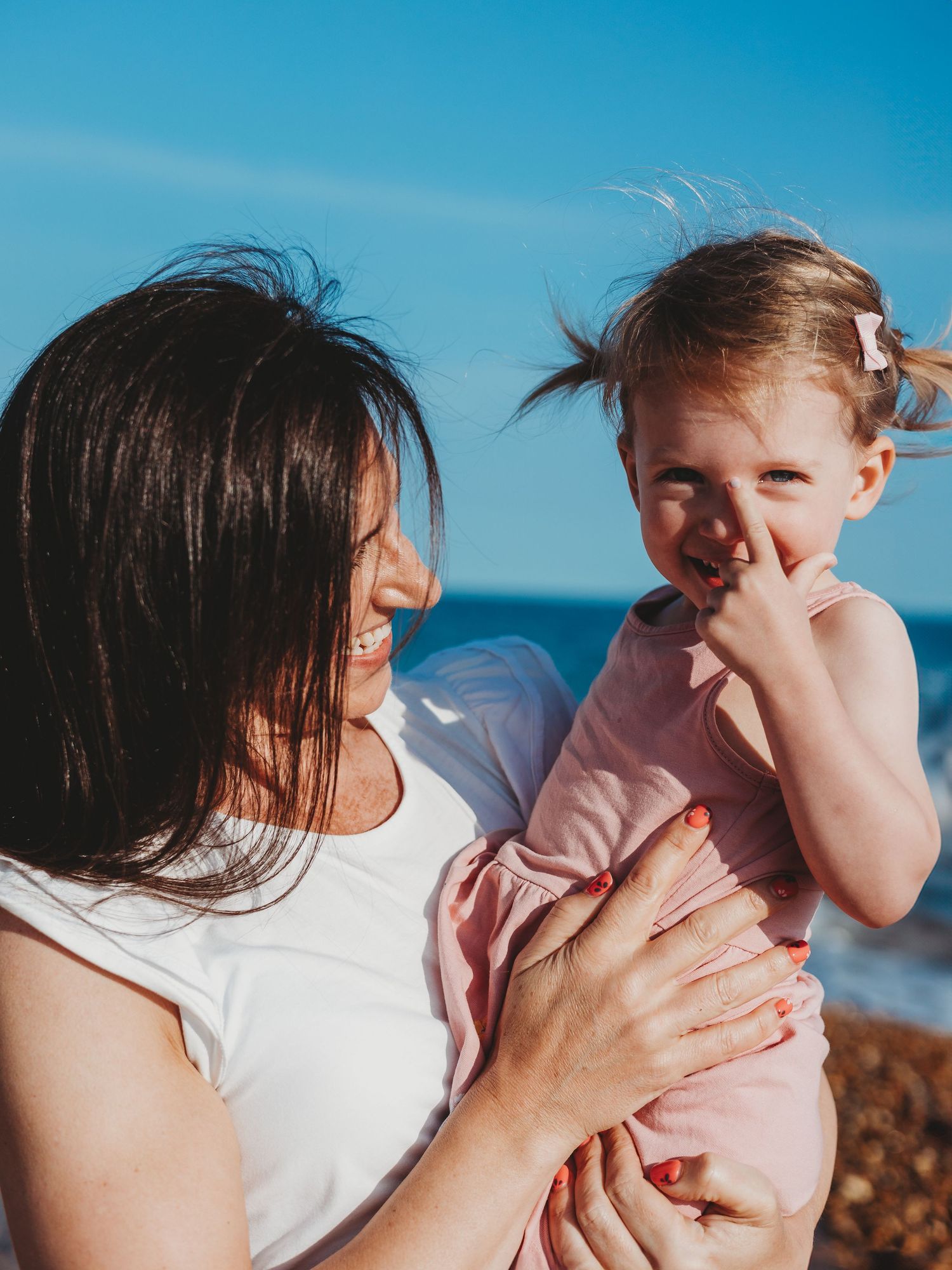 A smiling mother holds her laughing toddler girl in a pink dress at a sunny beach.